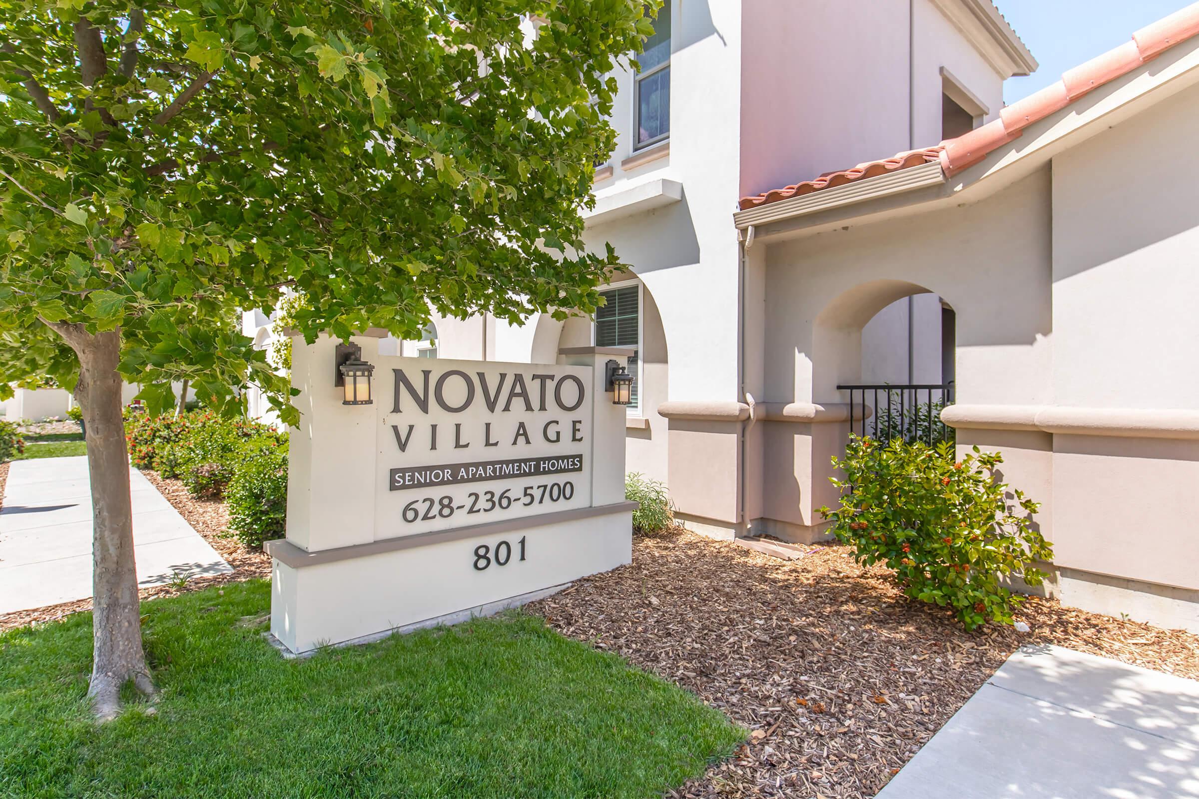 Signage for Novato Village Senior Apartment Homes, featuring the name "Novato Village" prominently displayed. The sign includes a contact number (628-236-5700) and the address "801." Surrounding the sign are landscaped areas with green grass and decorative materials, contributing to a welcoming atmosphere.