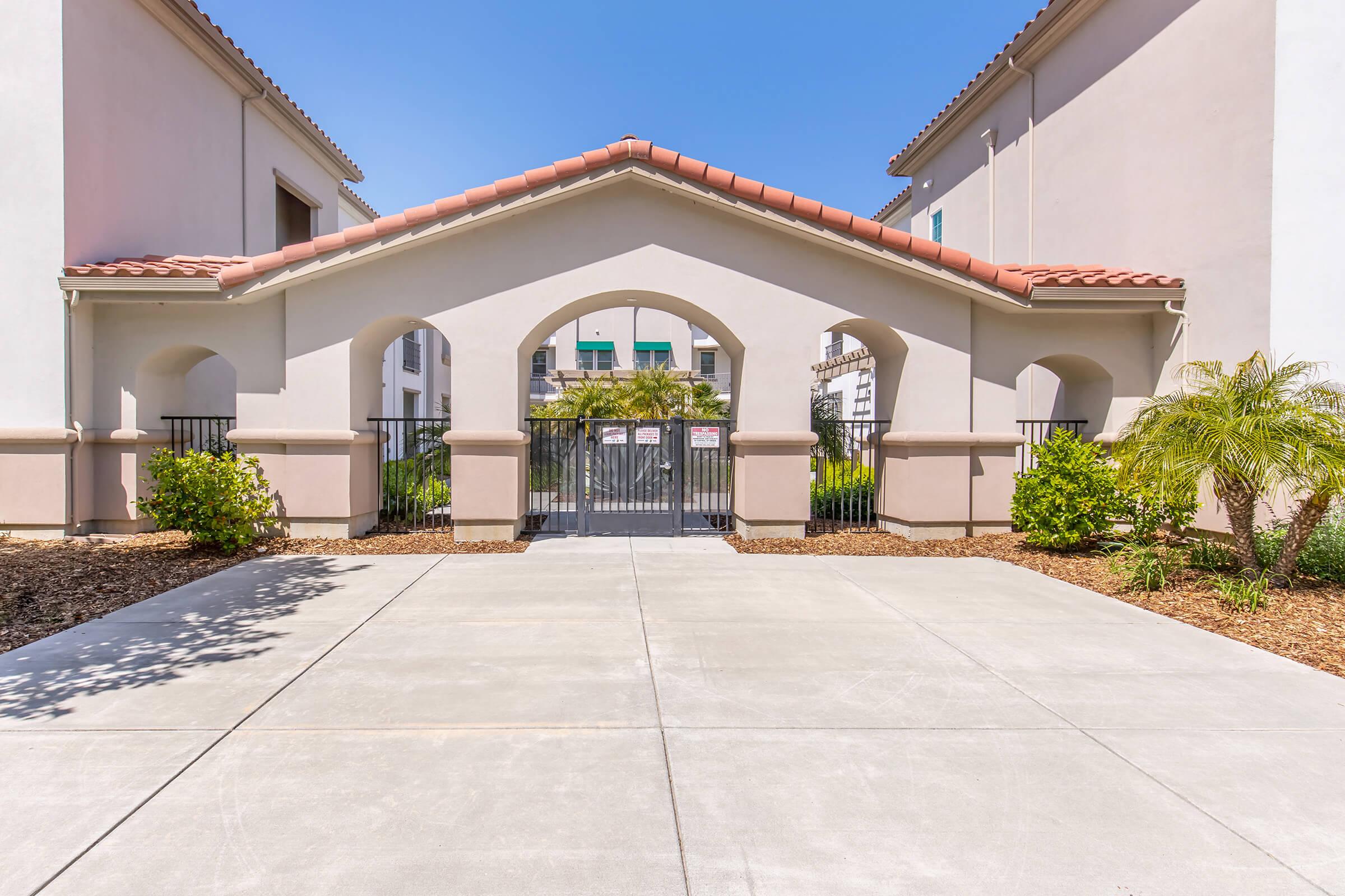 A gated entrance to a residential complex, featuring a tiled roof and arched entrance. The pathway is paved, with greenery and palm plants on either side. The buildings are painted in light colors, and a blue sky is visible above.