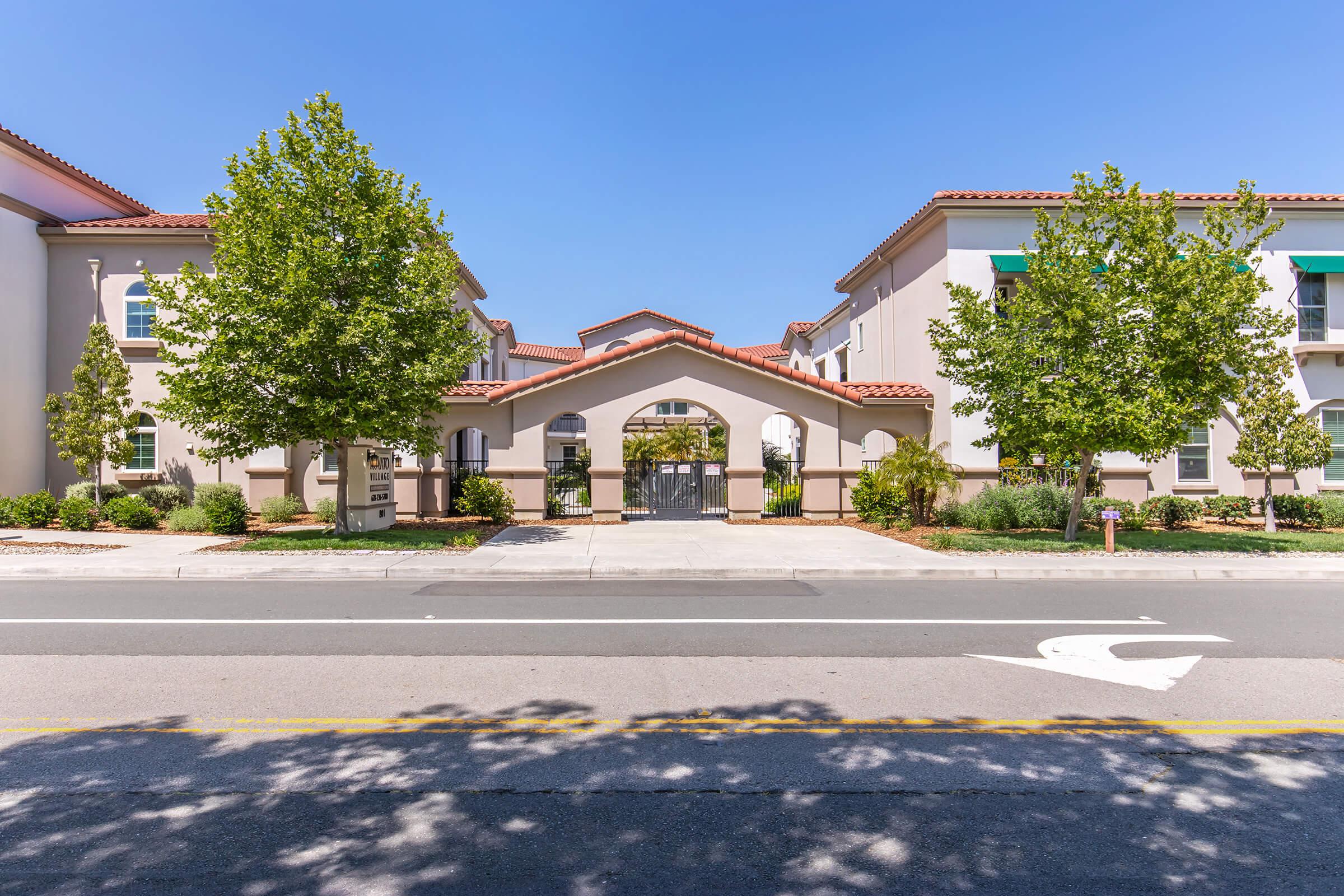 A view of a modern building featuring an arched entrance, flanked by trees. The architecture has a Mediterranean style with a tiled roof. The foreground includes a landscaped area along a street, showcasing clear blue skies and a peaceful atmosphere.