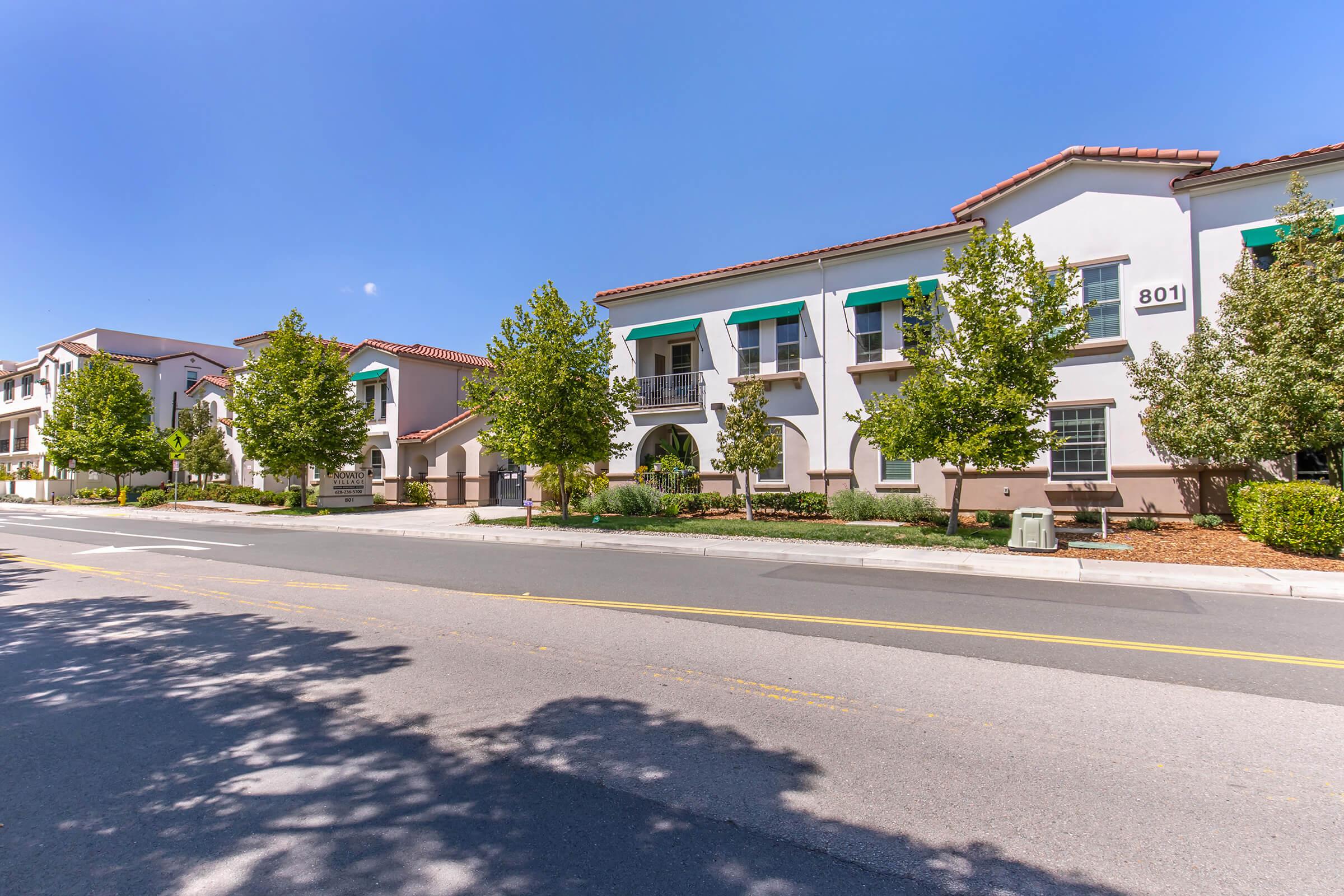 A quiet residential street featuring two-story apartment buildings with green awnings, surrounded by well-maintained trees and landscaping. The scene is set under a clear blue sky, showcasing a serene neighborhood atmosphere. A portion of the street is visible in the foreground.