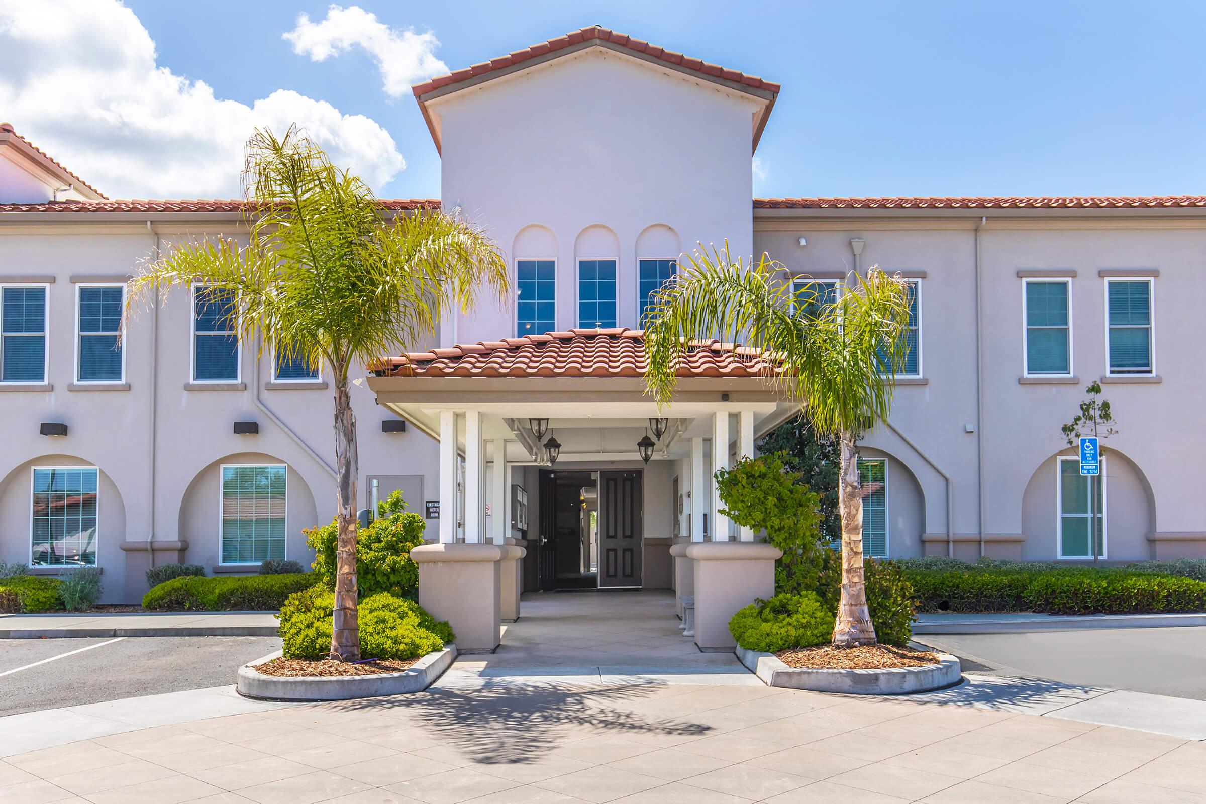A modern building with a light-colored exterior and a red-tiled roof. The entrance features tall columns and two palm trees on either side. The surrounding area contains neatly landscaped greenery and a paved driveway leading up to the entrance. Bright blue sky with a few clouds is visible.