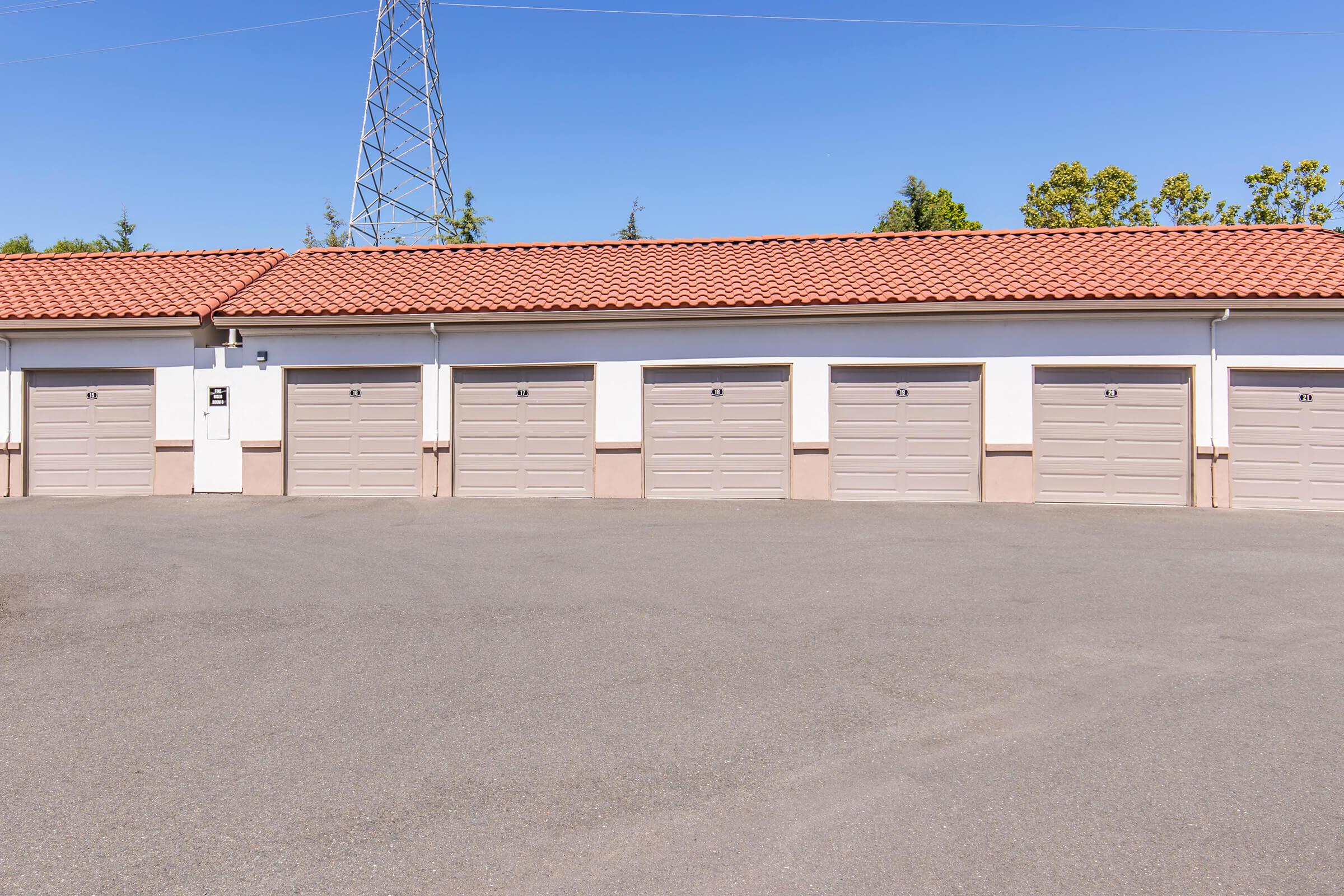 A row of closed garage doors with beige walls and a terracotta tiled roof. The area is paved with asphalt and a clear blue sky is visible above. A tall power line tower is seen in the background among trees.