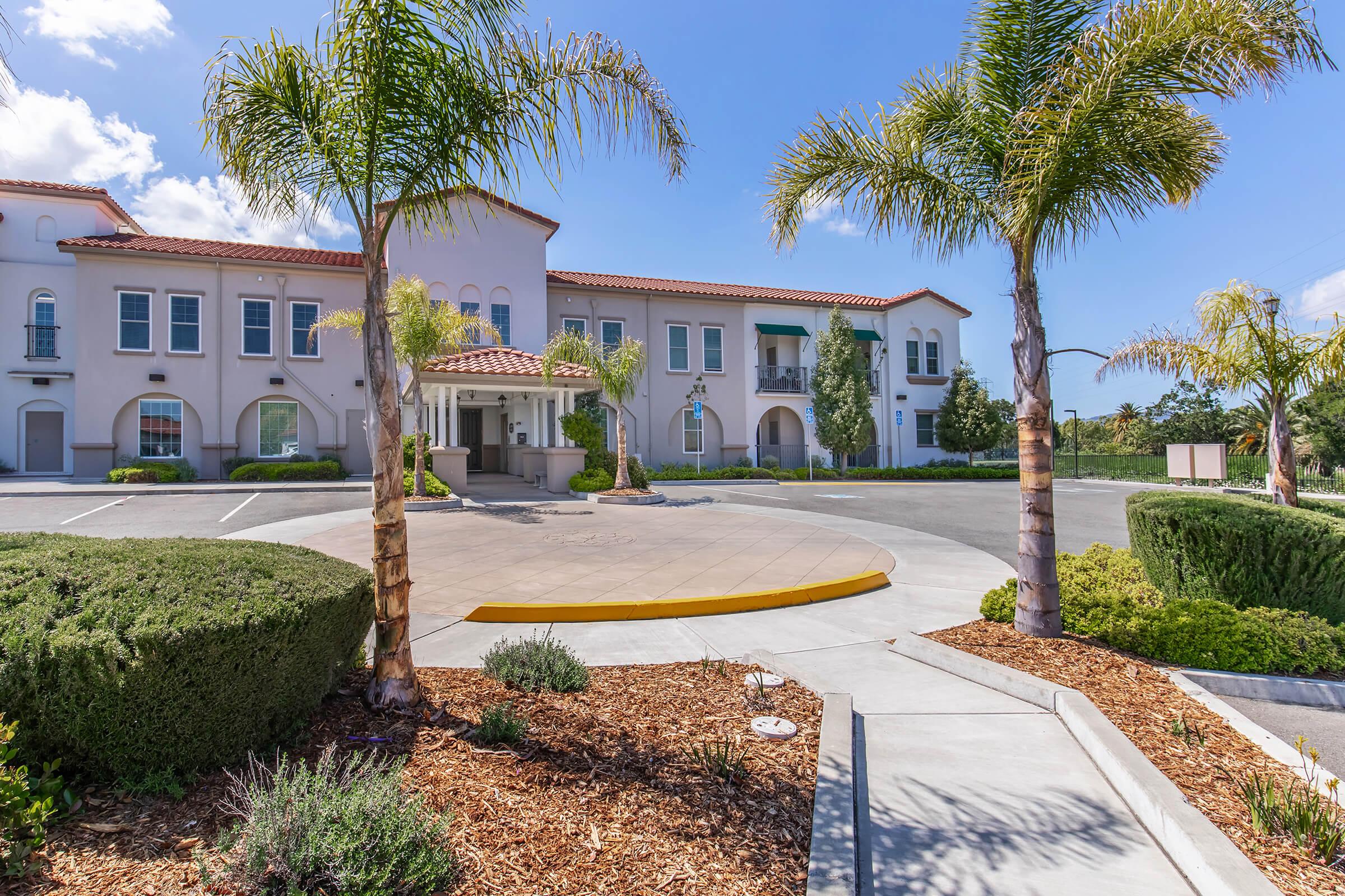 A modern building with a Mediterranean architectural style, featuring rounded arches, a red tile roof, and palm trees in the foreground. The entrance has a circular driveway, well-maintained landscaping with shrubs and mulch, and a clear blue sky in the background.