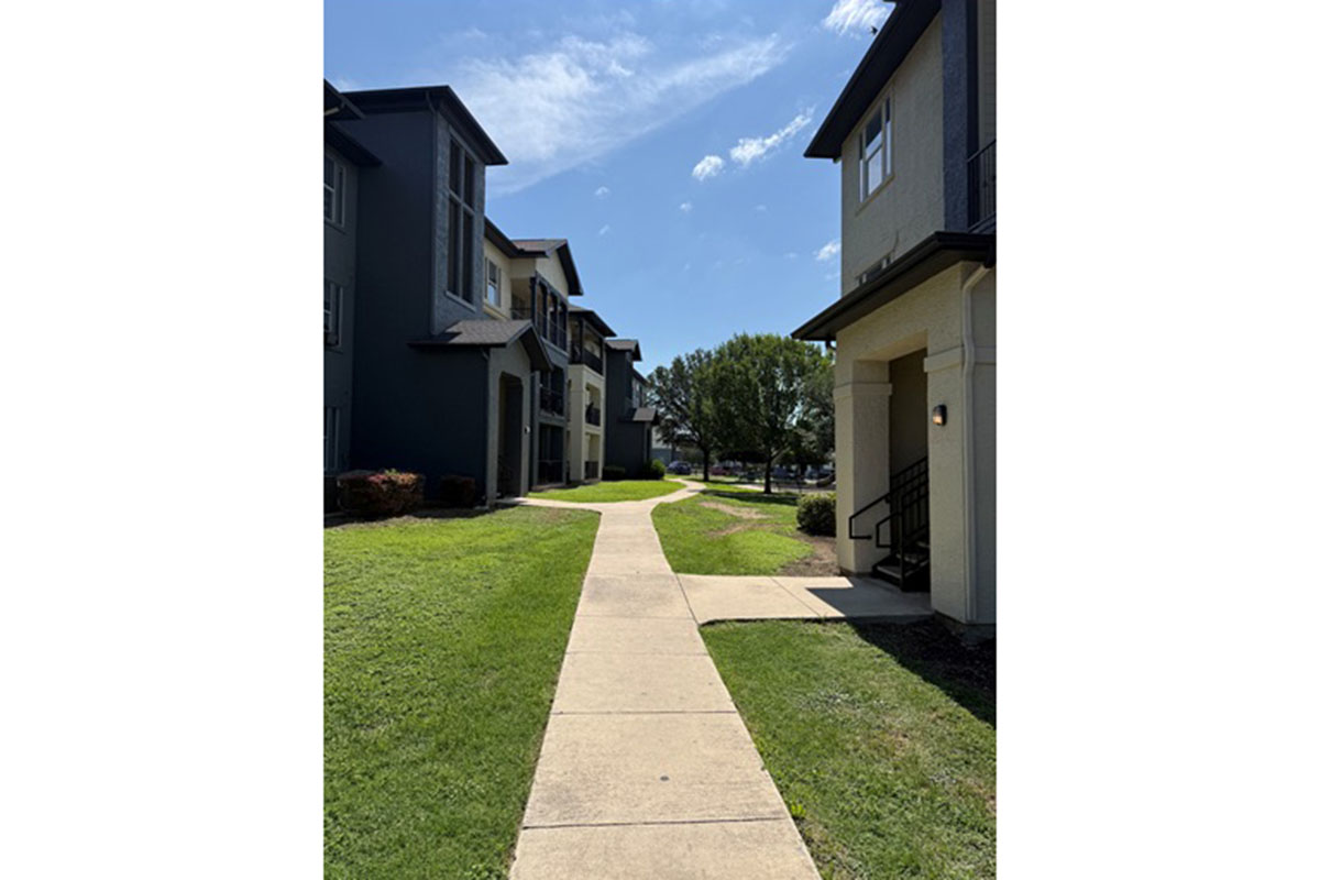 A pathway running between two multi-story apartment buildings, with well-maintained grass and trees on either side. The sky is clear with a few clouds, indicating a sunny day. One building features a staircase leading to an entrance.
