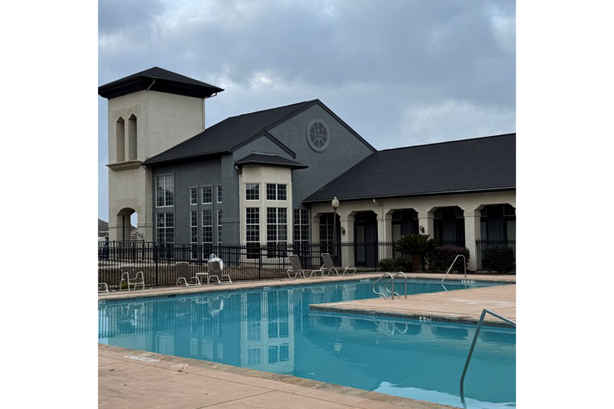 A modern building with a clock tower beside a swimming pool. The pool reflects the cloudy sky and is surrounded by a black fence. Lounge chairs are placed around the pool area, creating a relaxing atmosphere. The architecture features large windows and a combination of light and dark-colored exterior walls.