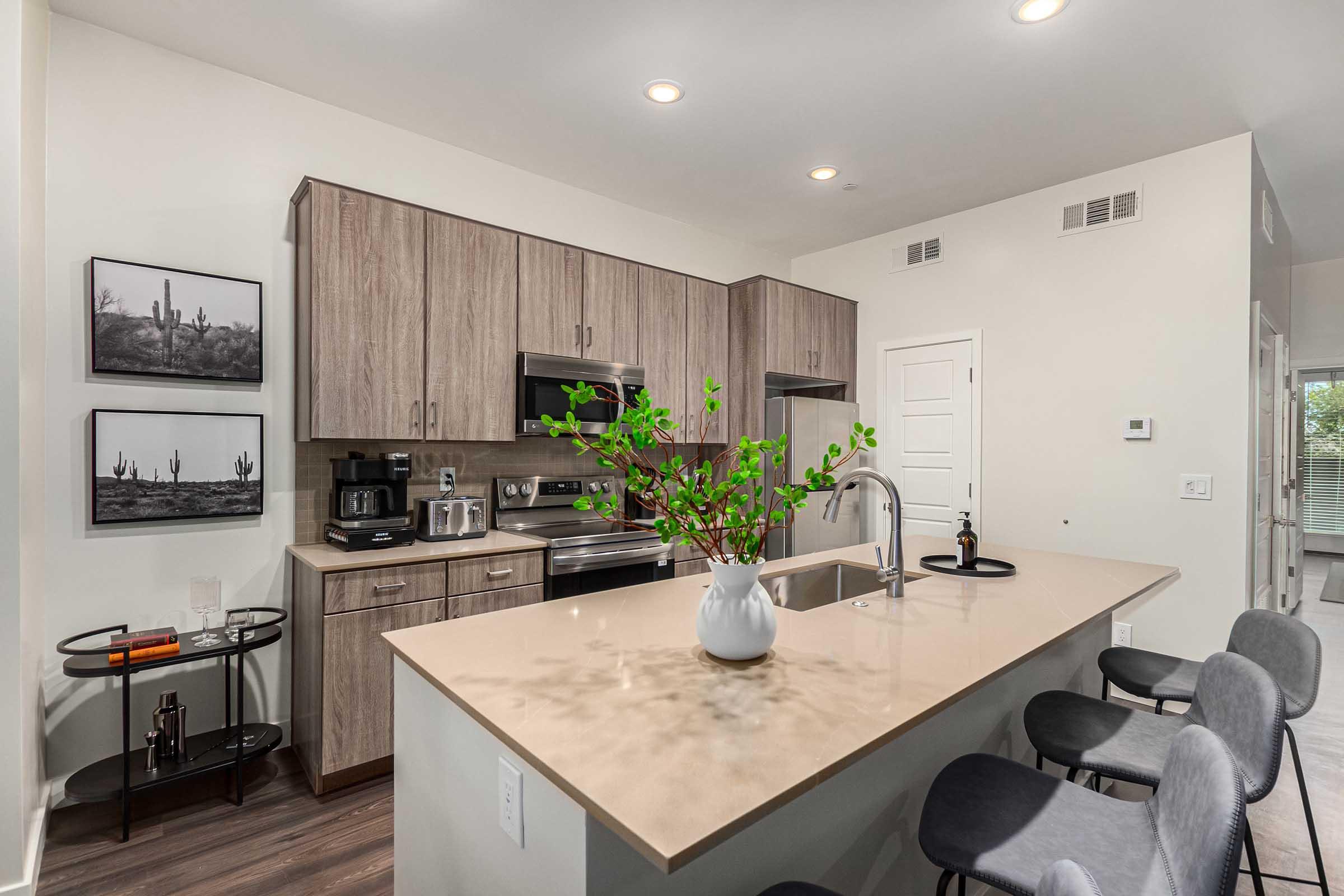 Modern kitchen featuring light wood cabinetry, stainless steel appliances, and a beige countertop. There are two framed black-and-white photographs of cacti on the walls. A small plant arrangement is on the counter, and a bar cart is positioned nearby with a few decorative items and a coffee machine.