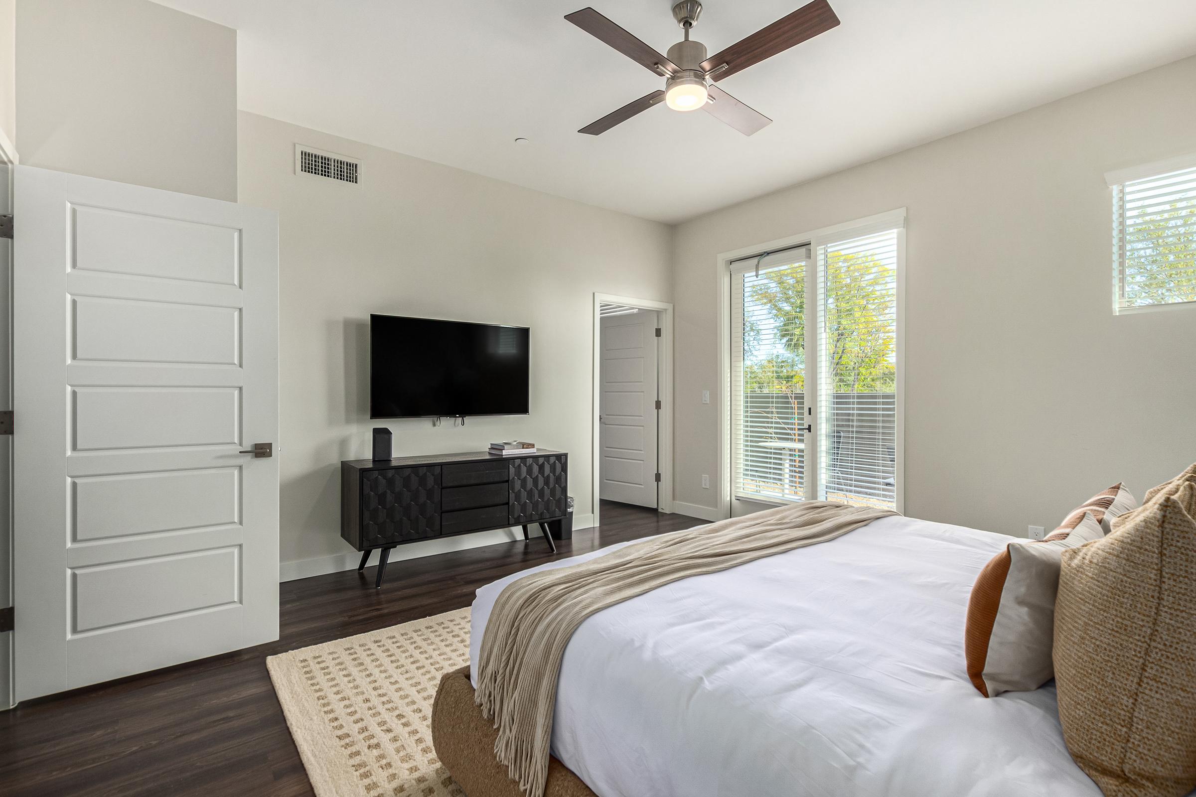 A modern bedroom featuring a white bed with a beige throw blanket, a dark wood dresser with a flat-screen TV above it, and large windows allowing natural light. The room has light-colored walls and dark wood flooring, complemented by a ceiling fan and a neutral color palette.