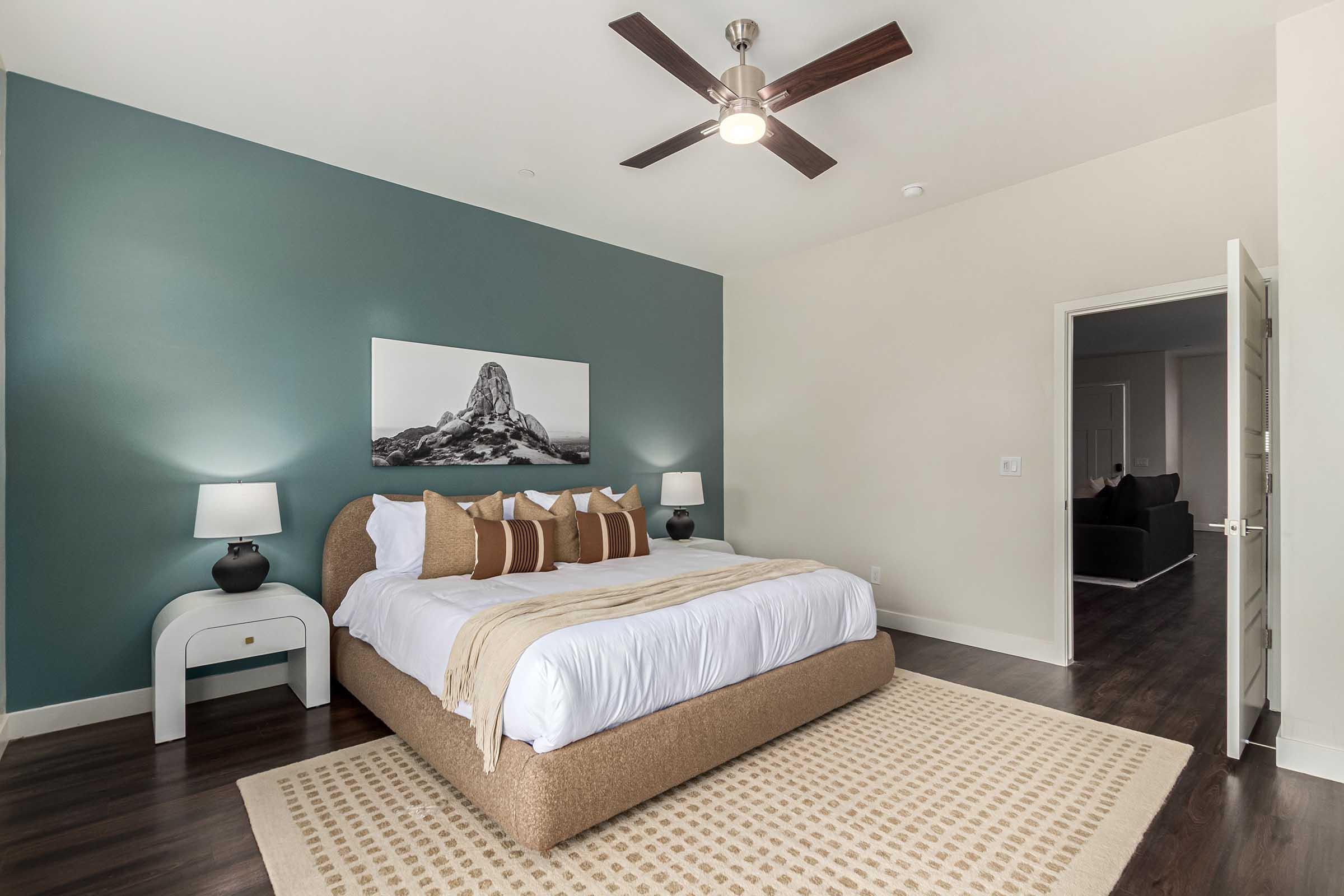 A modern bedroom featuring a large bed with white linens and decorative brown pillows. Two bedside lamps sit on either side, and a photo of a rocky landscape is mounted on the teal accent wall. The room has wooden flooring and a light-colored rug, creating a cozy atmosphere. A doorway leads to another room.