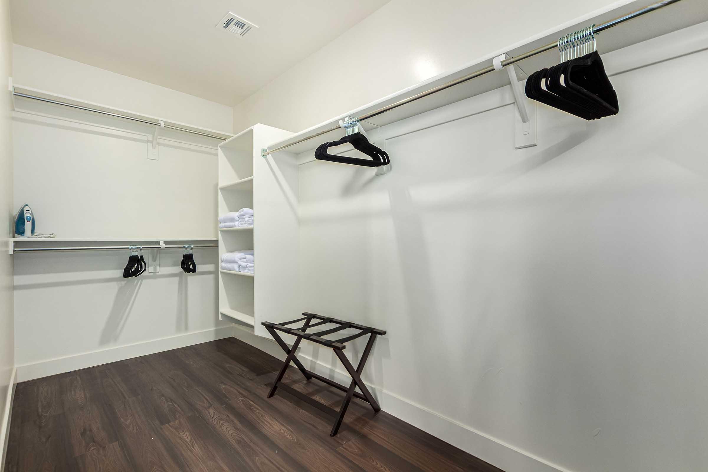 An empty walk-in closet featuring white walls and dark flooring. There are two hanging rods with black hangers, a small folding stool, and shelves displaying some rolled towels. The space is well-organized and minimalistic, ideal for storage or displaying clothing.