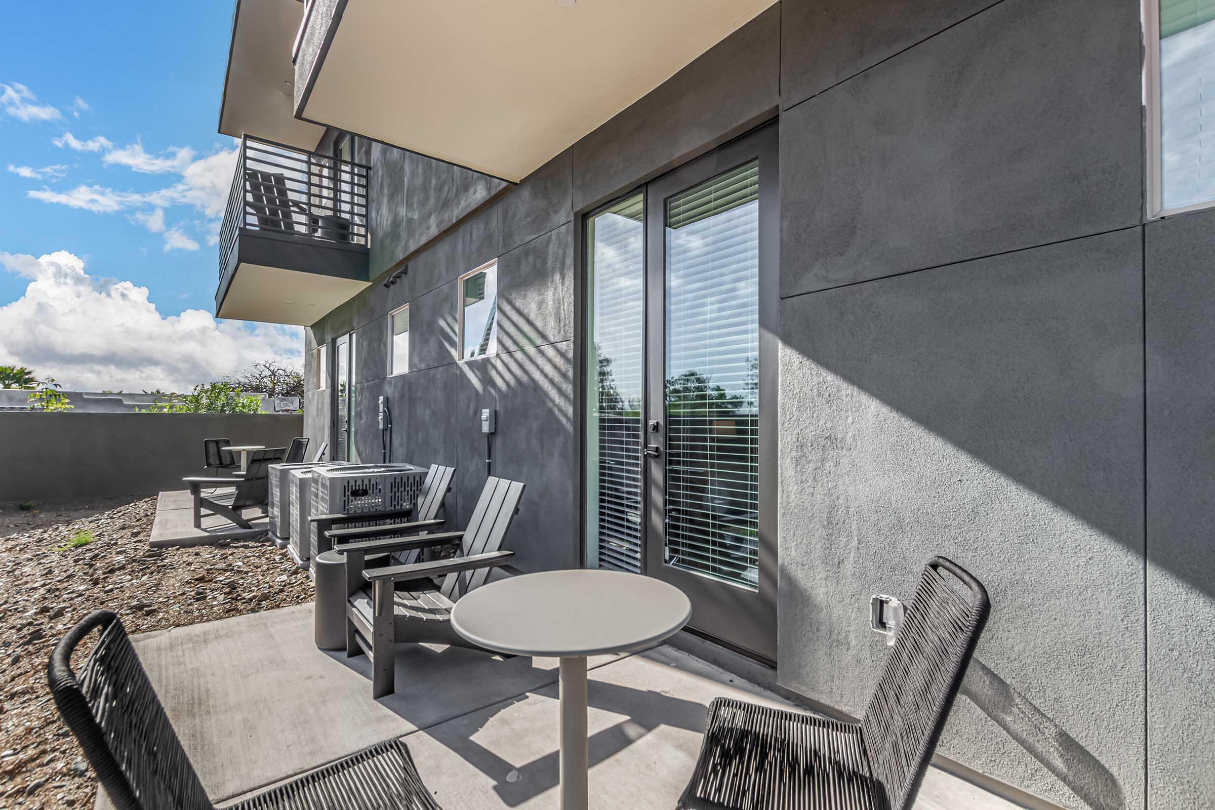 Outdoor patio area featuring a circular table and several black chairs. The setting includes a modern building with large windows, a gravel landscape, and blue skies peeking through scattered clouds.