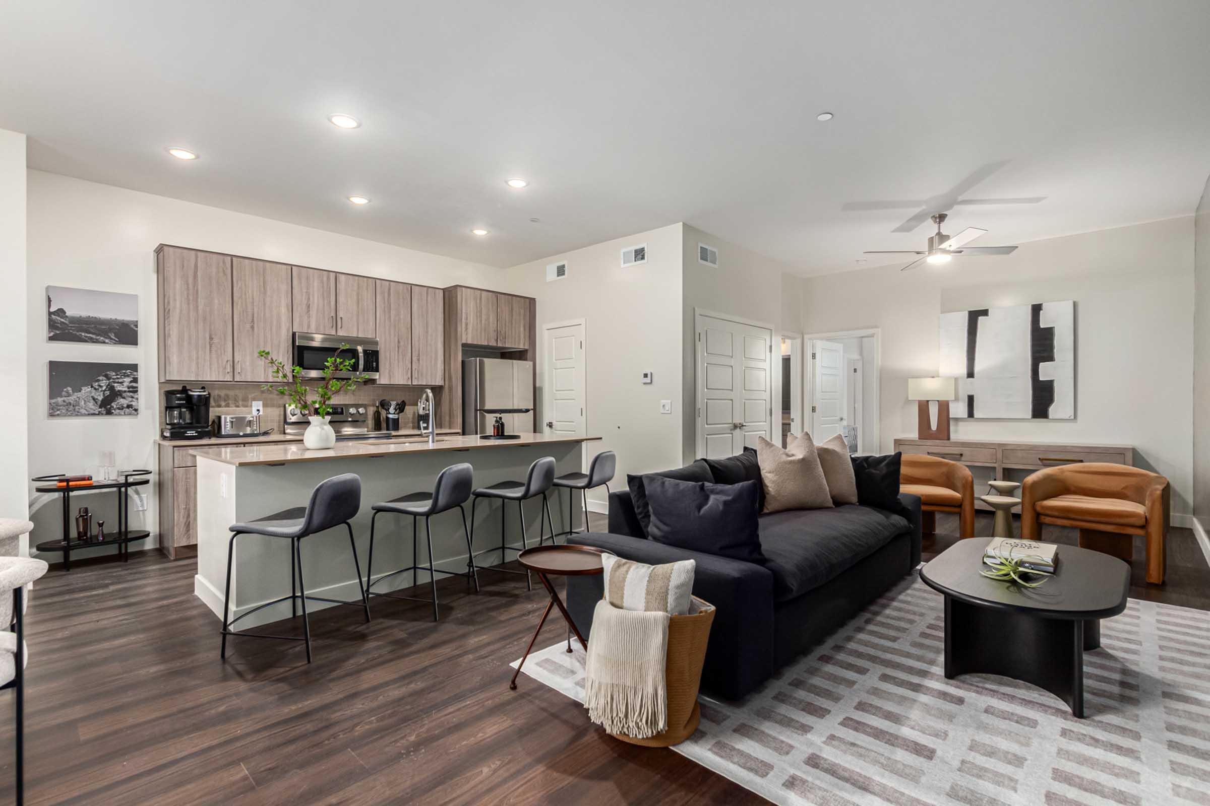 Modern open-concept living area featuring a dark sofa with decorative pillows, a sleek coffee table, and stylish chairs. The kitchen includes bar stools, wooden cabinetry, and appliances. Light-colored walls and hardwood flooring create a spacious ambiance, complemented by artistic wall décor and ample natural light.