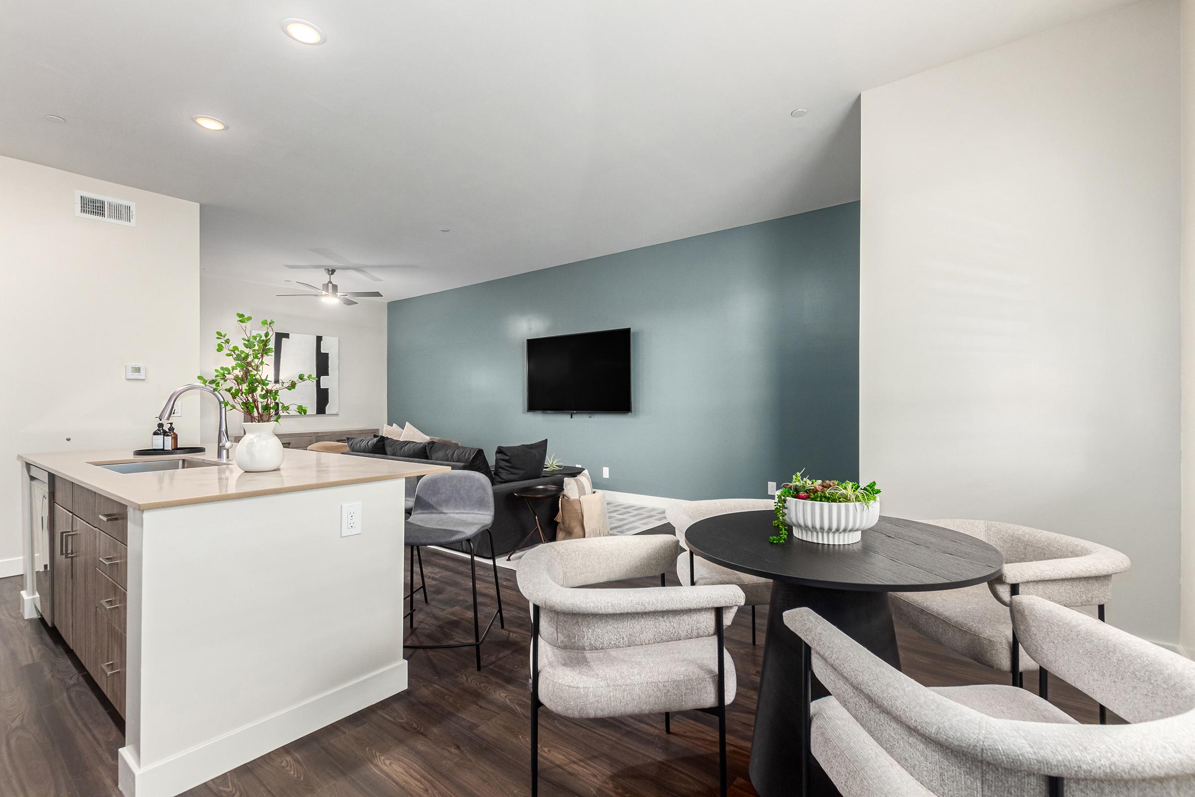 Modern open-concept living space featuring a kitchen with an island, a dining area with circular black table and chairs, and a cozy lounge area. There's a large television mounted on a teal accent wall, and neutral decor complements the wooden flooring. Natural light brightens the room.