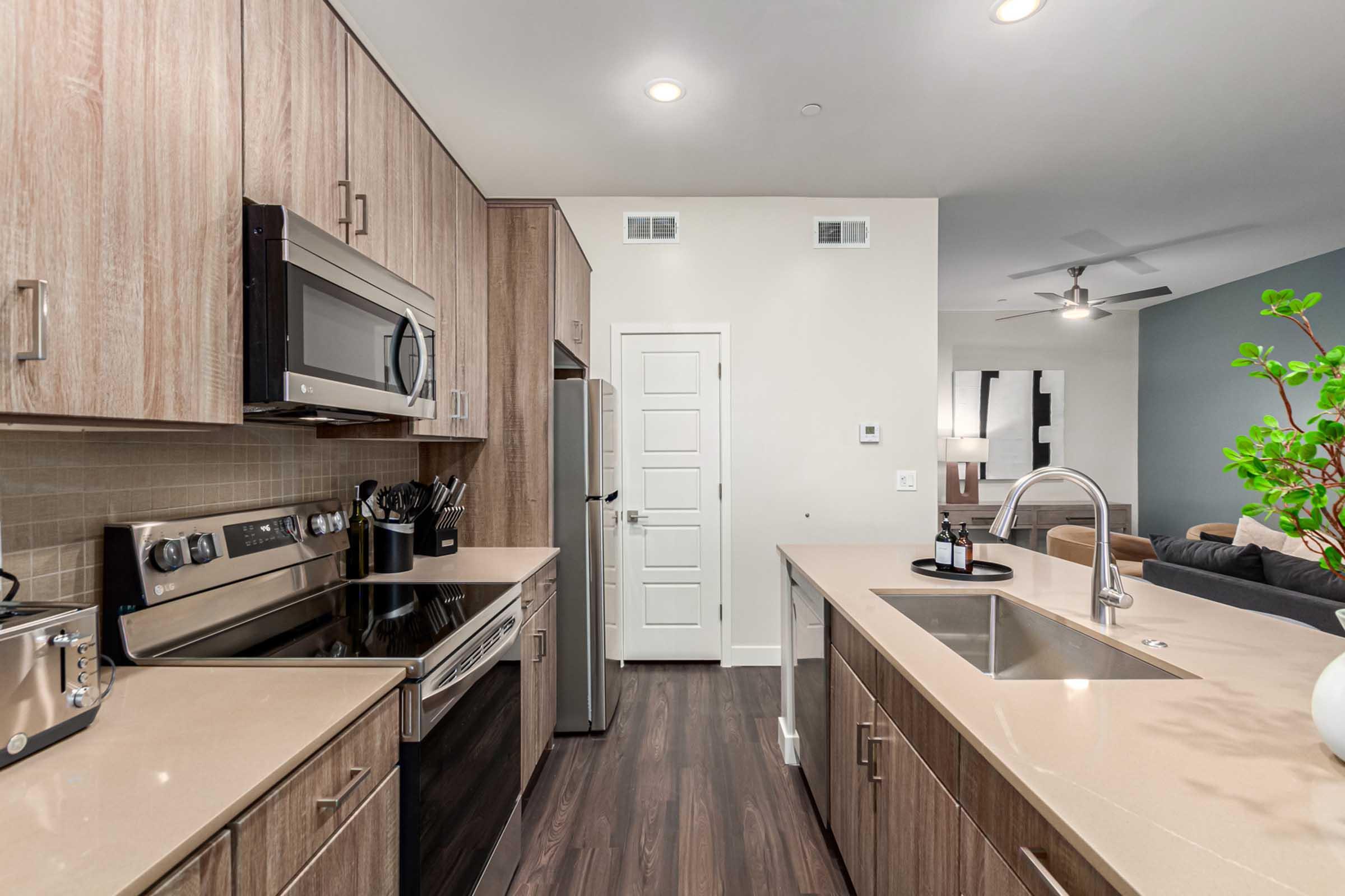 Modern kitchen featuring wooden cabinets, stainless steel appliances, and a sleek countertop. A sink is integrated into the kitchen island, and there is a door leading to another room. The walls are light-colored, and there is a plant on the countertop, enhancing the contemporary aesthetic.