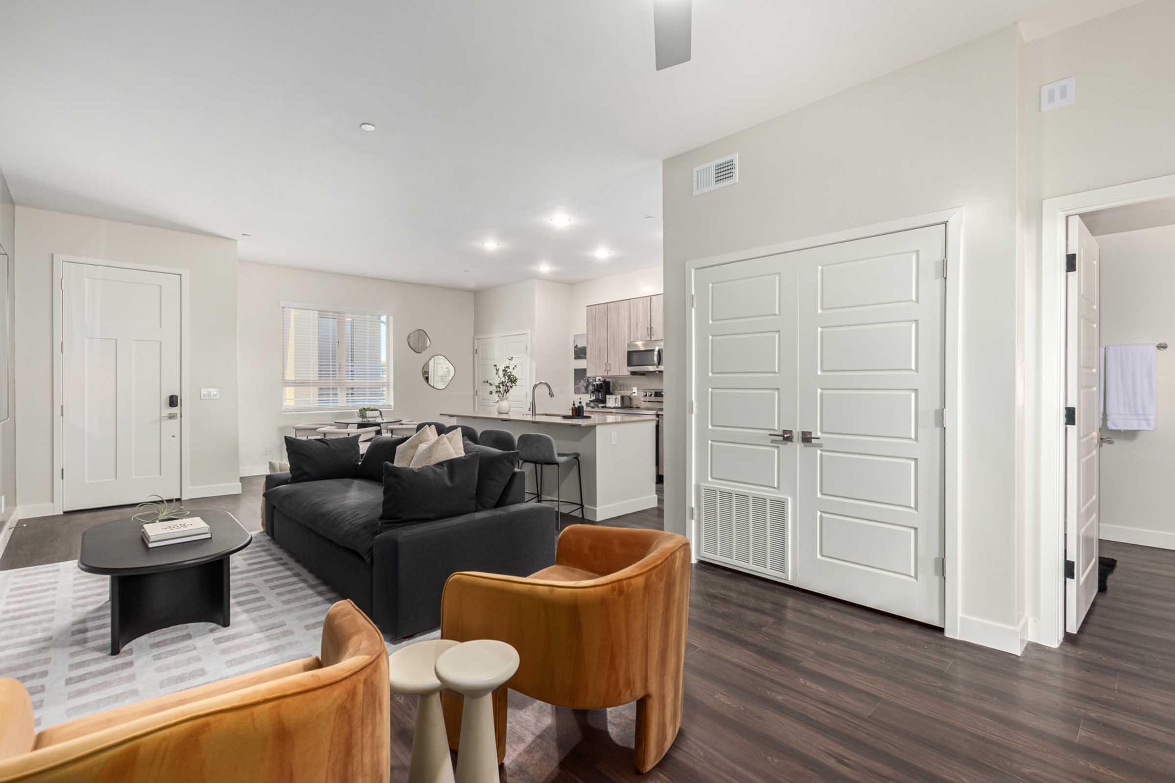 A modern living room and kitchen area featuring a black sofa, two orange chairs, and a coffee table. The space has light-colored walls, wooden flooring, and a bright ambiance, with large windows providing natural light. The kitchen is equipped with contemporary appliances and cabinets.