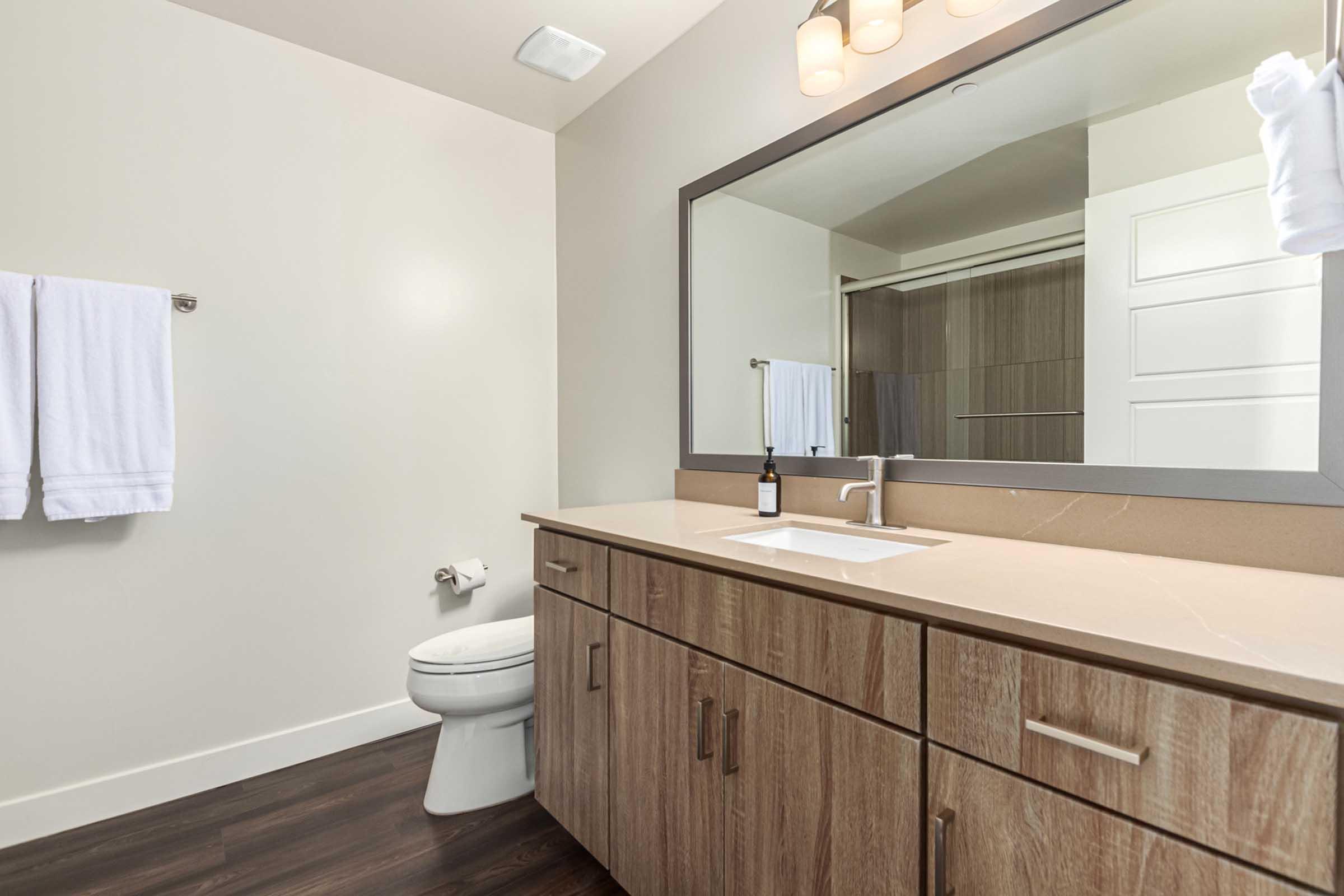 A modern bathroom featuring a wooden vanity with a sink, a large mirror above it, and two white towels hanging on a rack. There's a toilet in the foreground, and the shower area is visible in the background. The floor has dark wood-like tiles, creating a contemporary aesthetic.