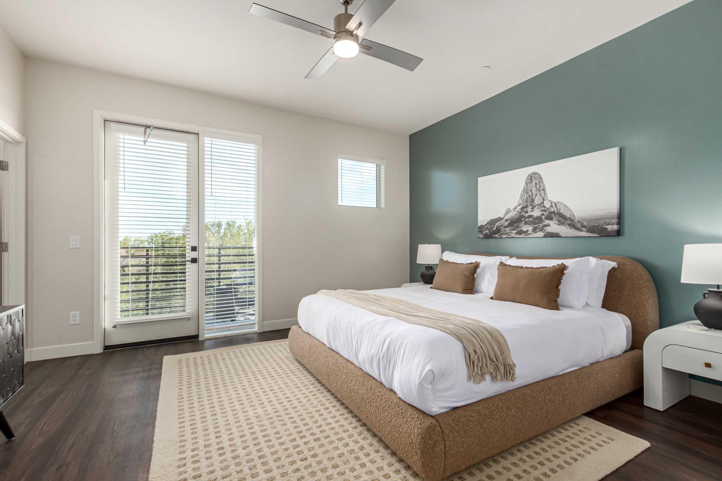 A modern bedroom featuring a plush bed with brown accent pillows and a neutral throw blanket. There are two bedside lamps on either side, a large window with blinds, and a balcony door providing natural light. A mountain-themed artwork hangs on the wall above the bed, and a rug is laid on the dark wooden floor.