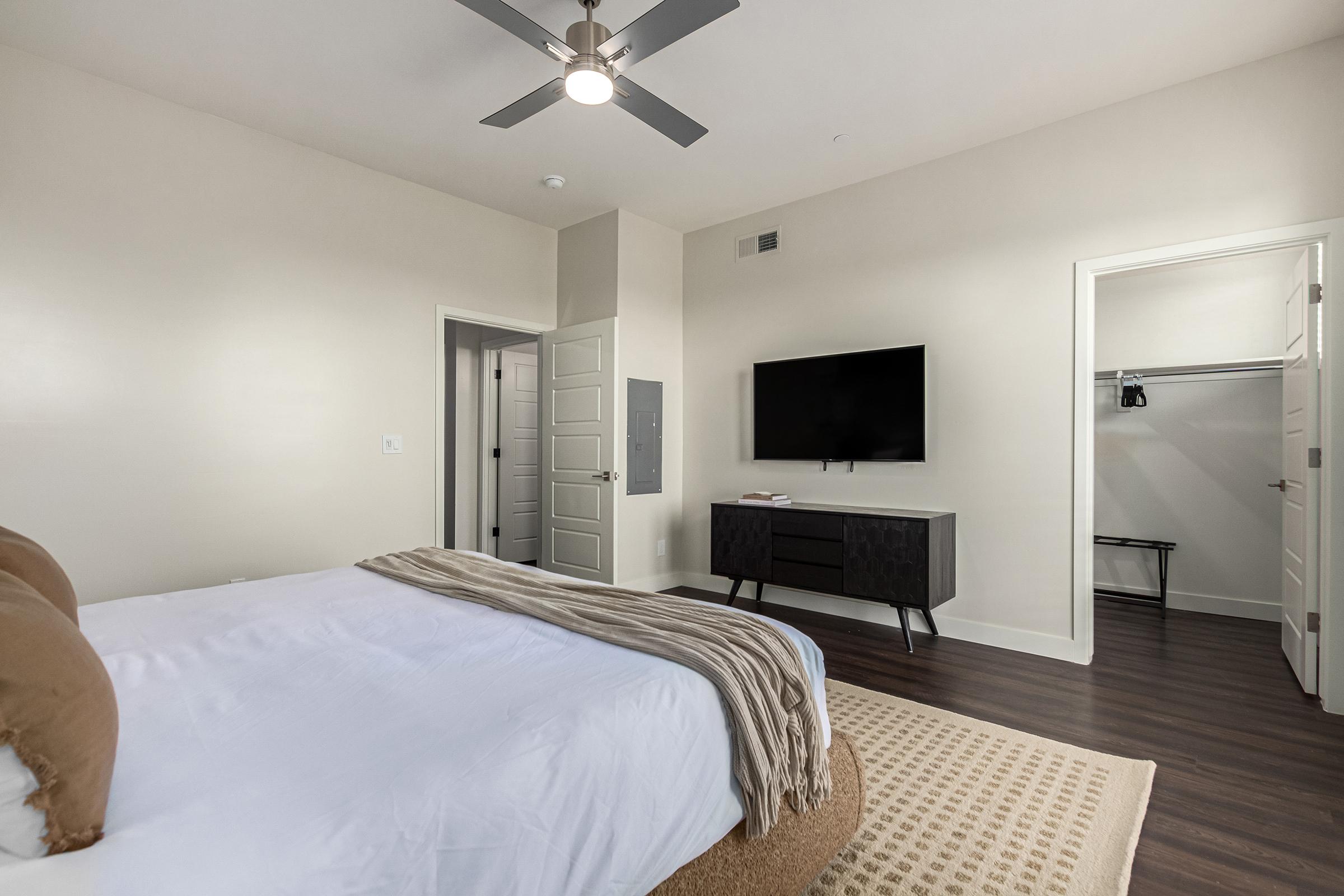A modern bedroom featuring a large bed with a brown throw blanket, a ceiling fan, a flat-screen TV mounted on the wall, a stylish dresser, and an open closet space. The walls are painted a light color, and the flooring is dark wood. A door leads to another room.