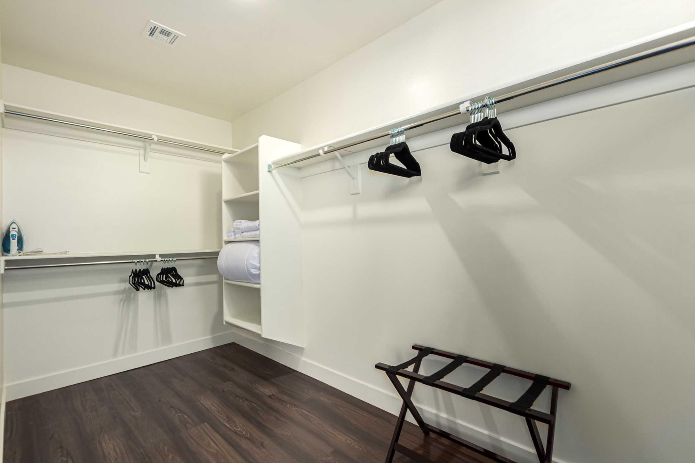 A spacious, minimalist closet featuring white walls, dark wooden flooring, and a combination of hanging rods with black hangers, a shelf with neatly rolled towels, an iron on a shelf, and a wooden folding clothes rack.