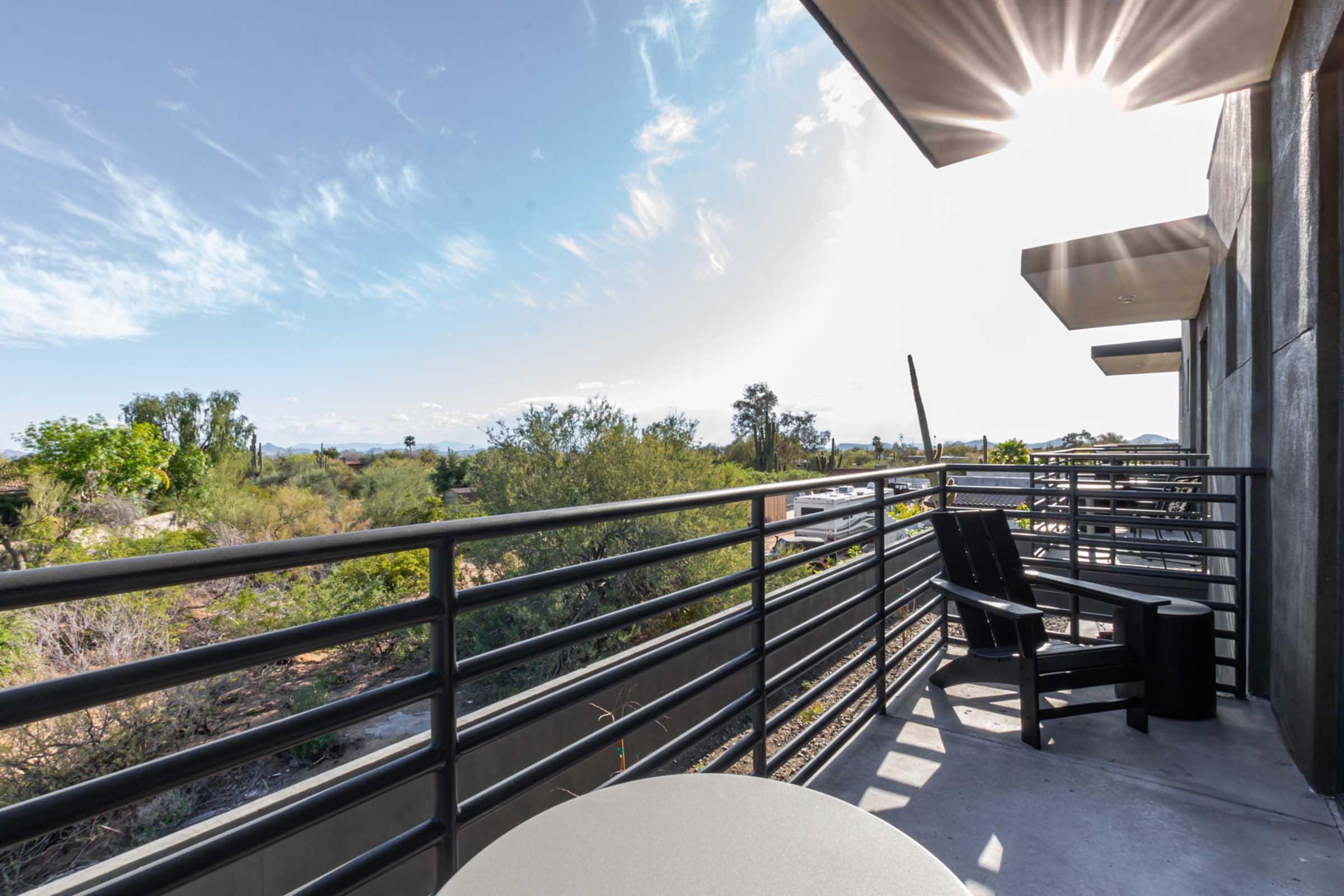 A spacious balcony with a table and chairs overlooks a green landscape with trees and shrubs. The sky is bright with a few clouds, and sunlight shines through the structures above, creating a warm atmosphere. The scene captures a blend of nature and modern living.
