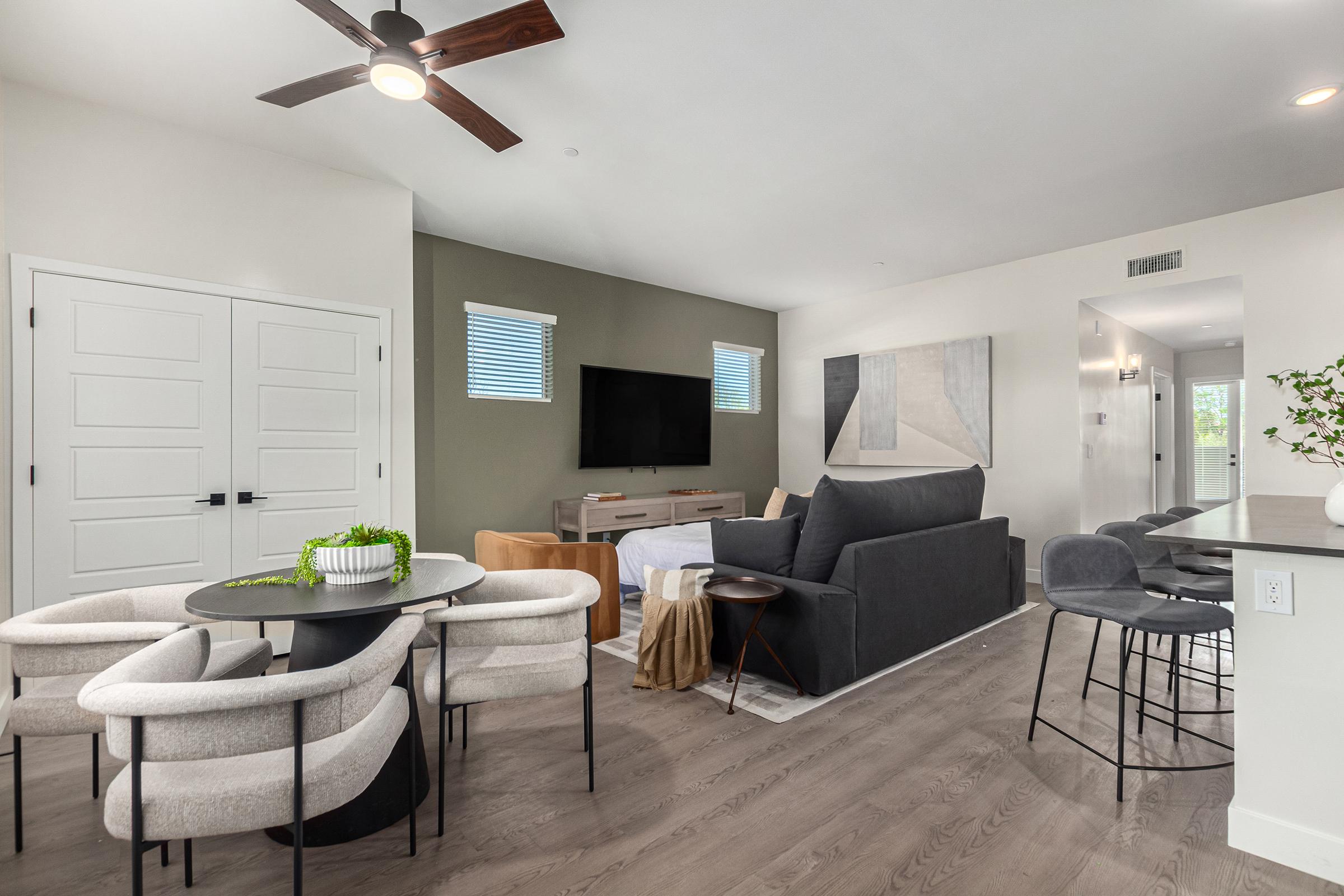 A modern living space featuring a dark sofa, a round dining table with plush chairs, and a wall-mounted TV. The room has light-colored walls, hardwood flooring, and large windows. Decorative elements include a small plant and an abstract art piece, creating a stylish and inviting atmosphere.
