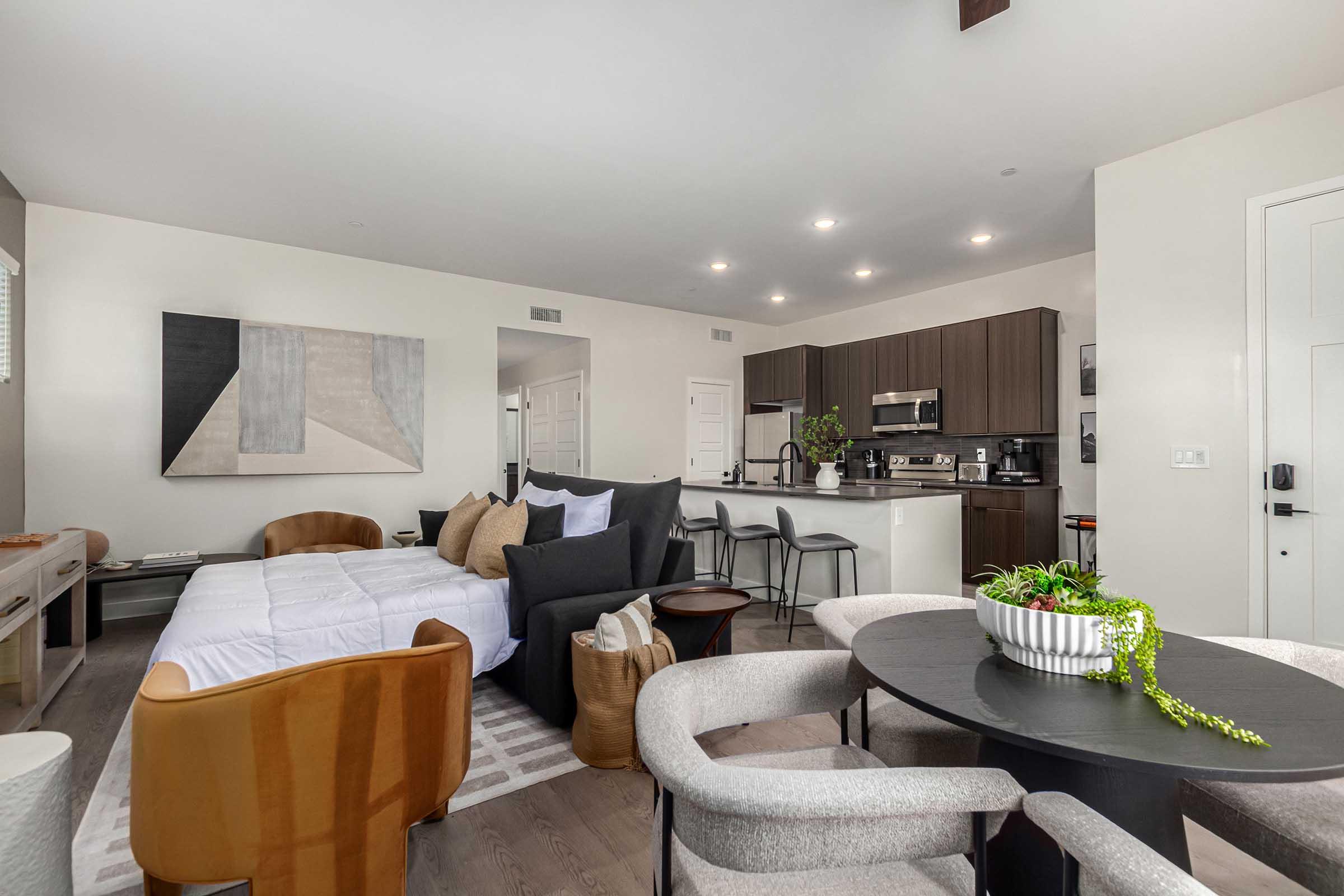 A modern studio apartment featuring a cozy seating area with a black sofa, decorative pillows, and a round dining table. The kitchen, equipped with dark cabinets, is visible in the background, along with stylish decor and plants enhancing the contemporary aesthetic. Soft lighting creates a warm atmosphere.