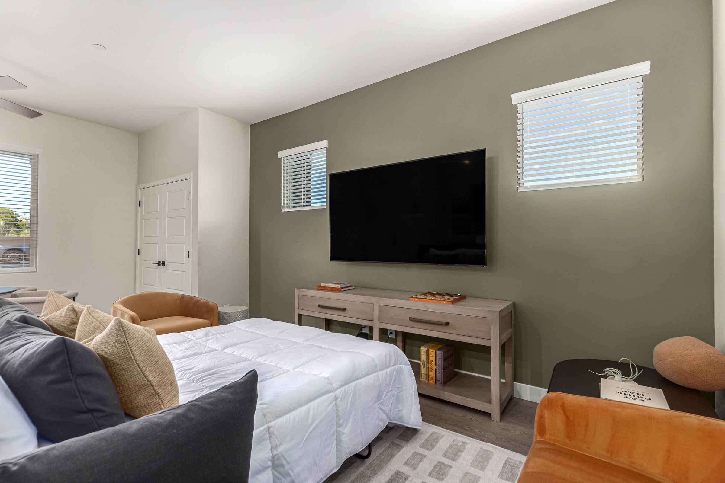 A modern living space featuring a wall-mounted television above a light wooden console. The room has a bed with white bedding and a mix of decorative pillows. Two windows with blinds allow natural light to enter, and there is an orange armchair in the corner, complementing the neutral color scheme of the walls.