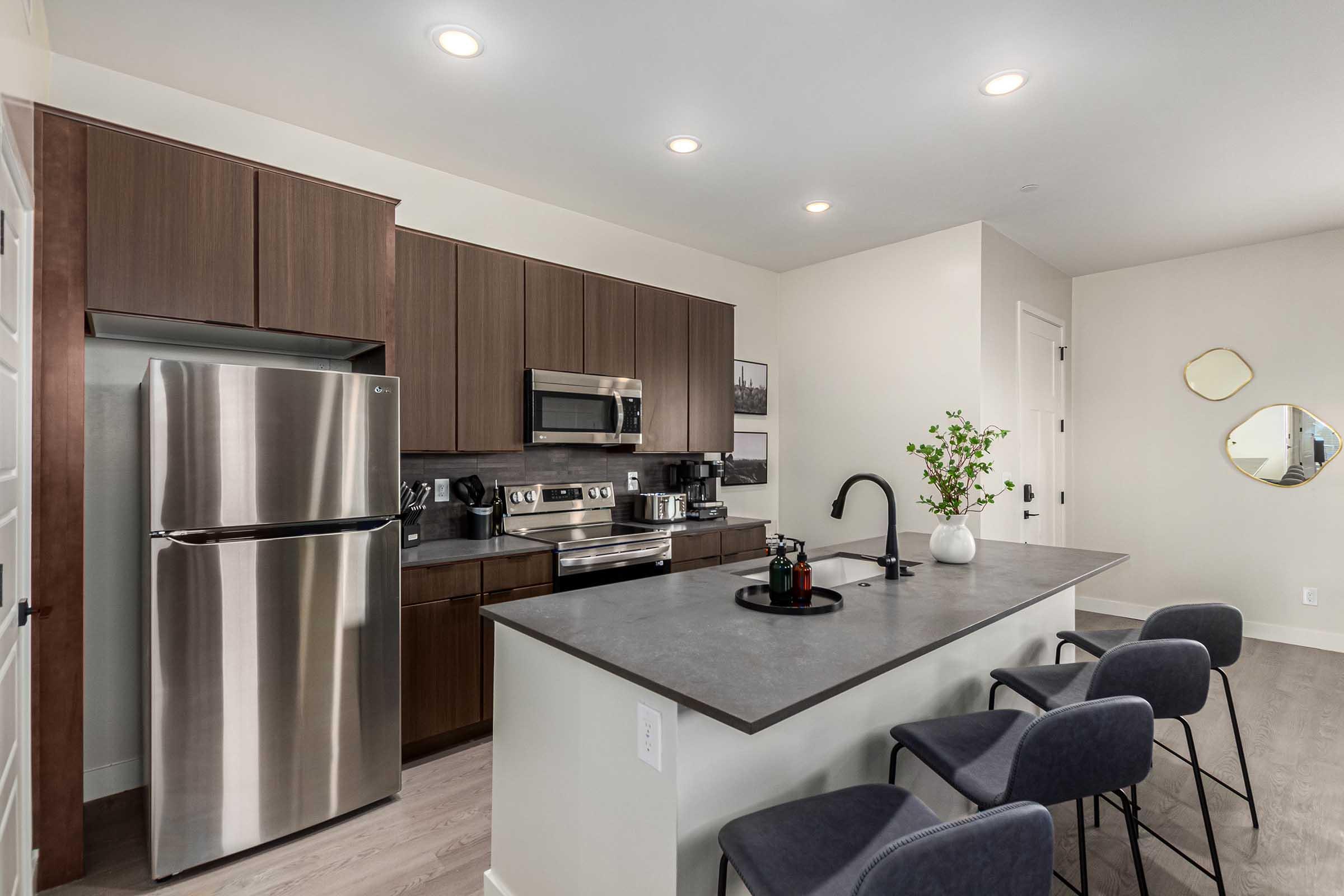 Modern kitchen featuring dark wood cabinetry, stainless steel appliances, and a gray island countertop. Bar stools are arranged around the island, and there is a small plant centerpiece. Bright, recessed lighting illuminates the space, creating a clean and inviting atmosphere.