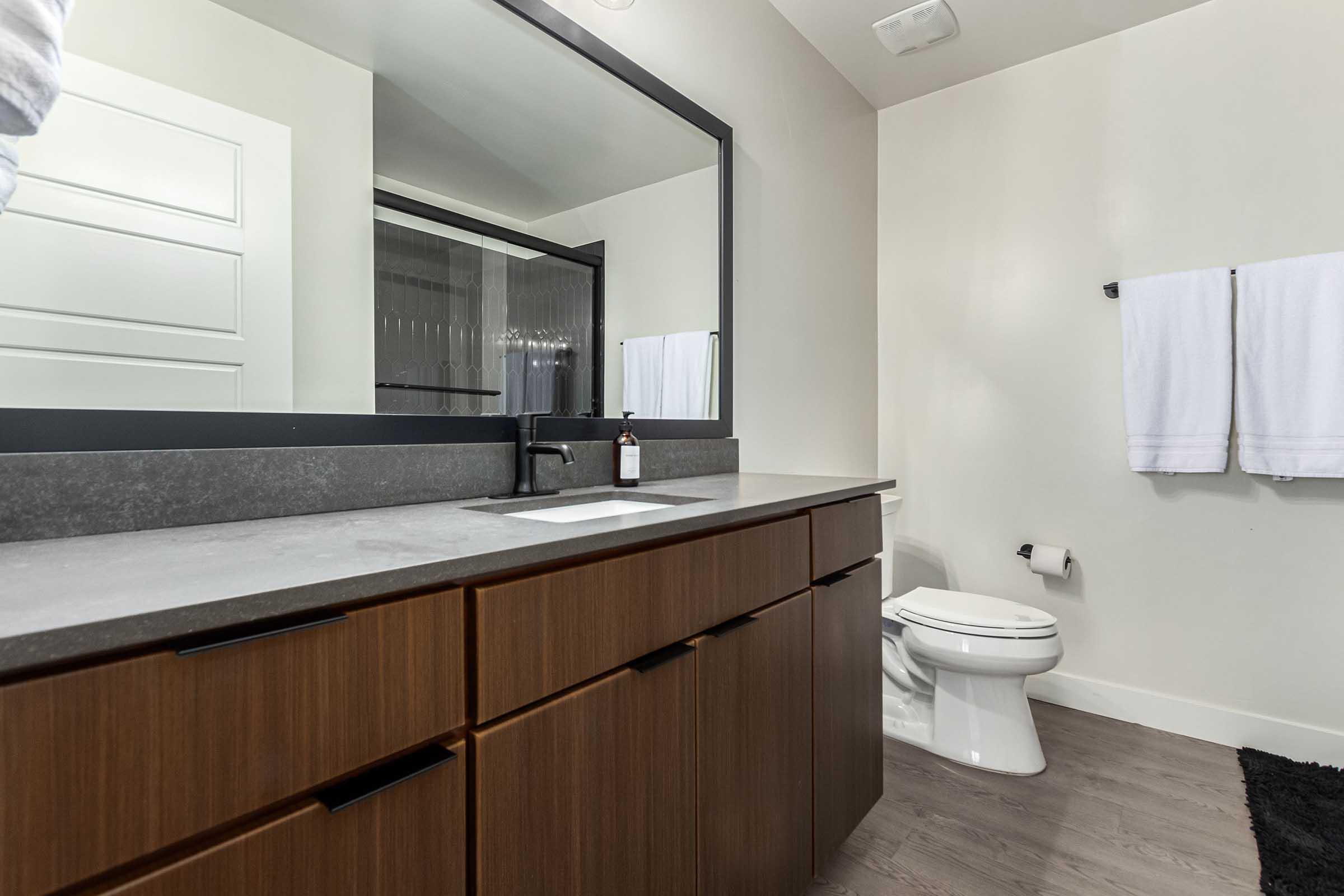 A modern bathroom featuring a sleek countertop with a sink, a large mirror, wooden cabinets, and a white toilet. Fresh towels hang on a rack, and a decorative black rug is on the floor. The overall design is clean and minimalist, showcasing neutral tones and stylish fixtures.