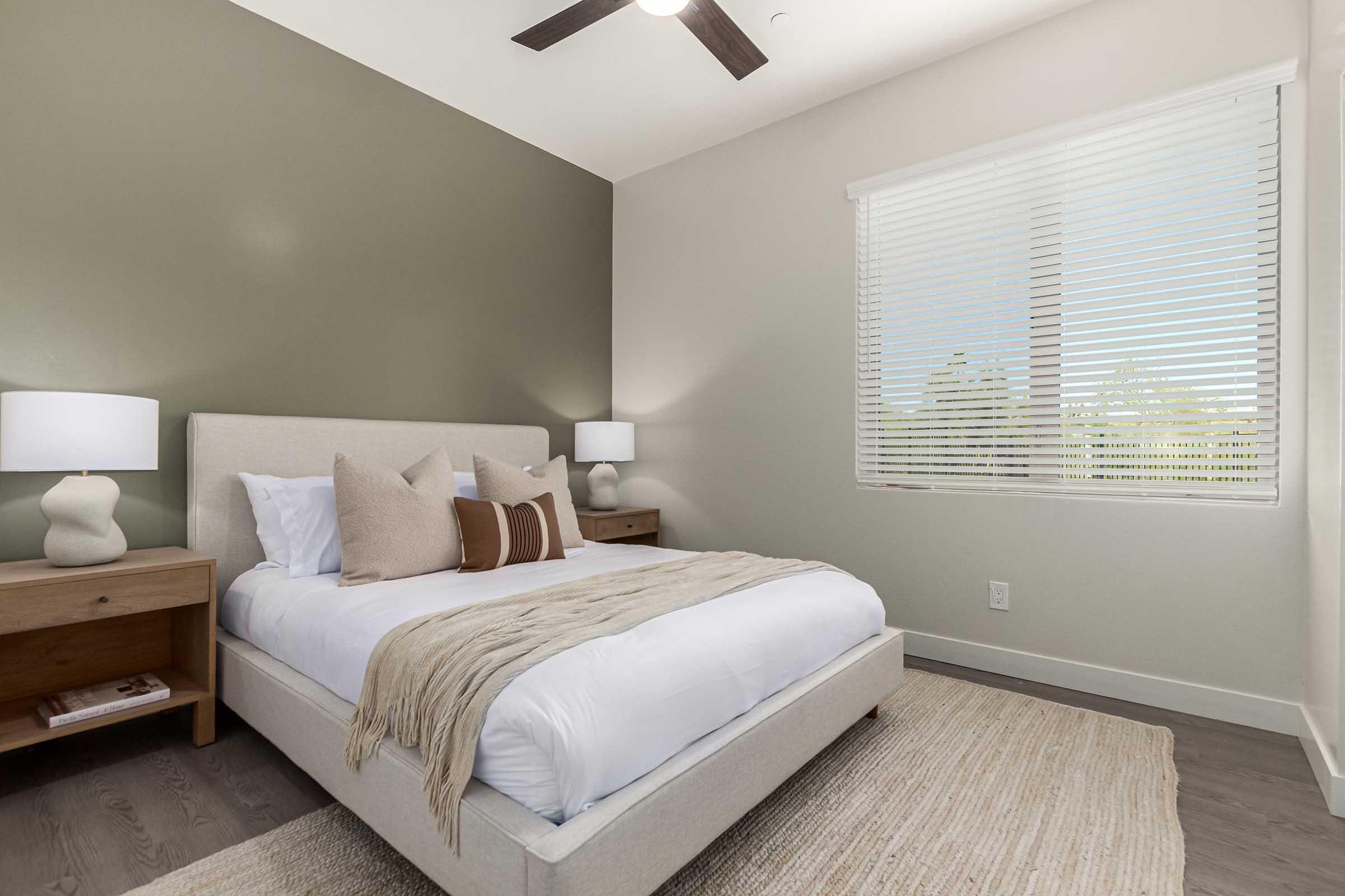 A modern bedroom featuring a light-colored upholstered bed with decorative pillows, two wooden nightstands with lamps, and a textured area rug. The walls are a soft green and beige, and there are large windows with blinds allowing natural light to illuminate the space.