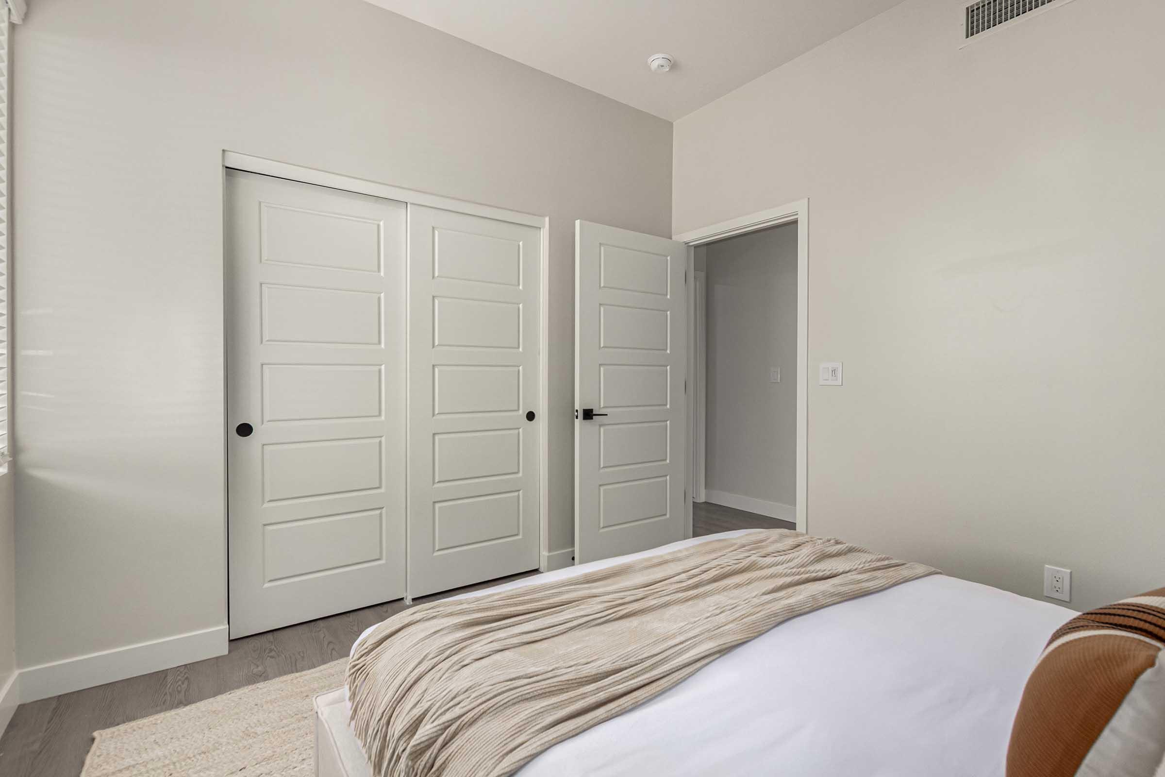 A well-lit bedroom featuring a bed with a light-colored blanket, a neutral wall color, and two closed doors leading to closets. The flooring is light wood, and there is a soft rug on the floor, creating a cozy and modern atmosphere. The room has a minimalistic design with no additional furnishings visible.