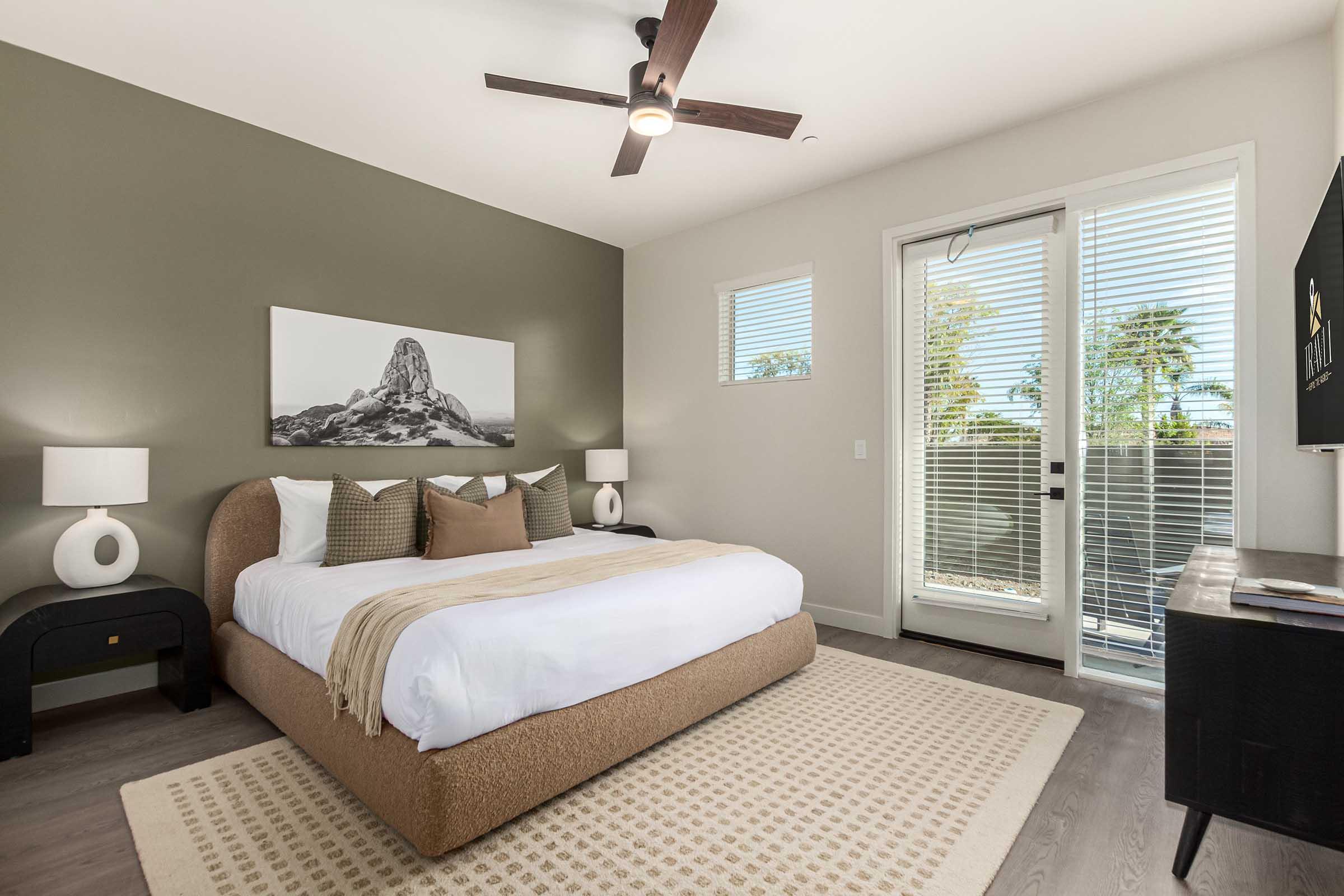 A modern bedroom featuring a large bed with a beige upholstered headboard, decorative pillows, and a light-colored throw blanket. There are two white lamps on nightstands, a minimalist black dresser, and a large window with blinds that opens to an outdoor view with palm trees. A monochrome mountain picture decorates the wall.