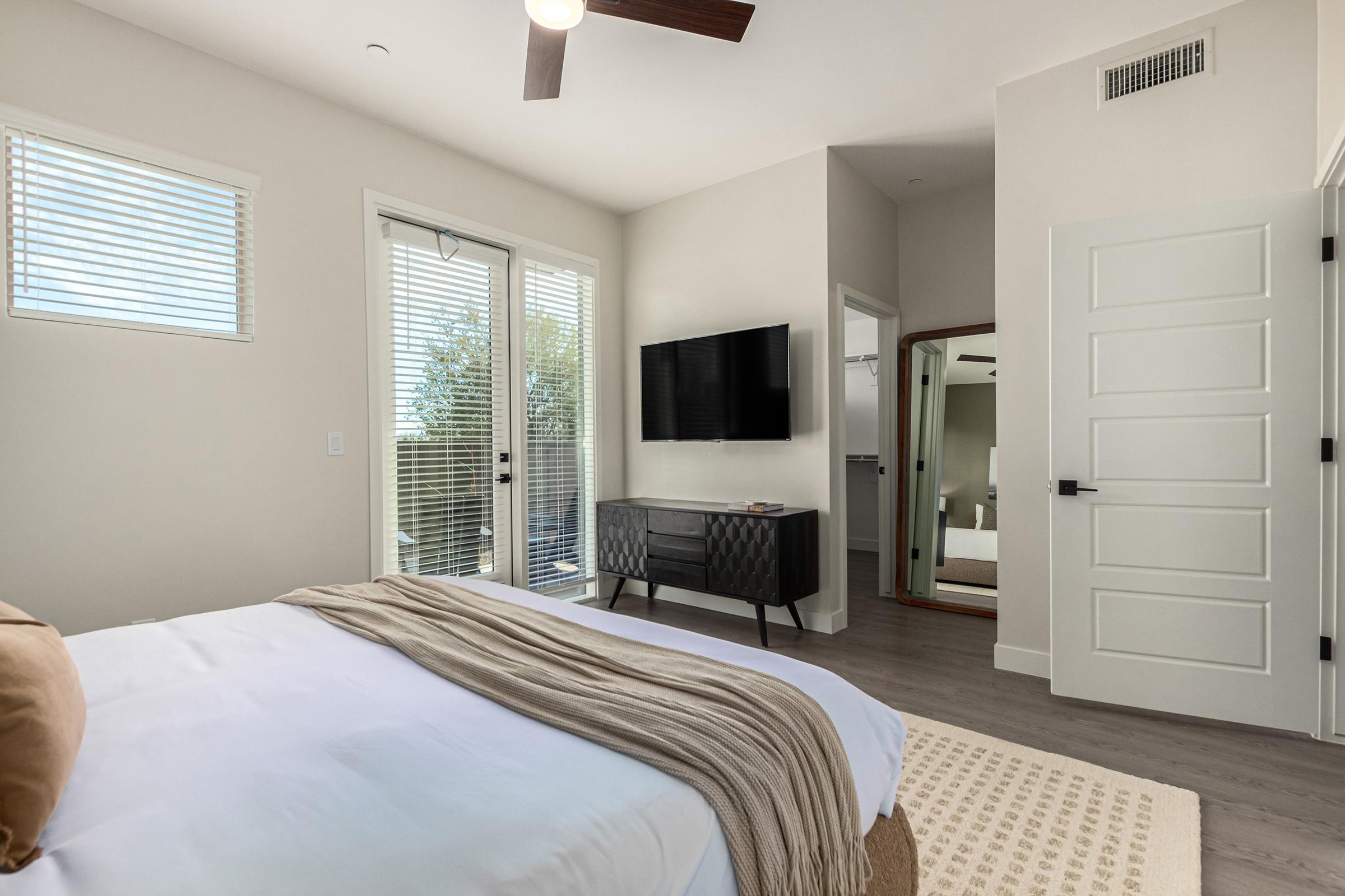 A modern bedroom featuring a bed with a beige throw, a nightstand, and a large window with blinds. There's a flat-screen TV mounted on the wall, a stylish dresser, and a mirror reflecting part of the room. Natural light fills the space, creating a warm and inviting atmosphere.