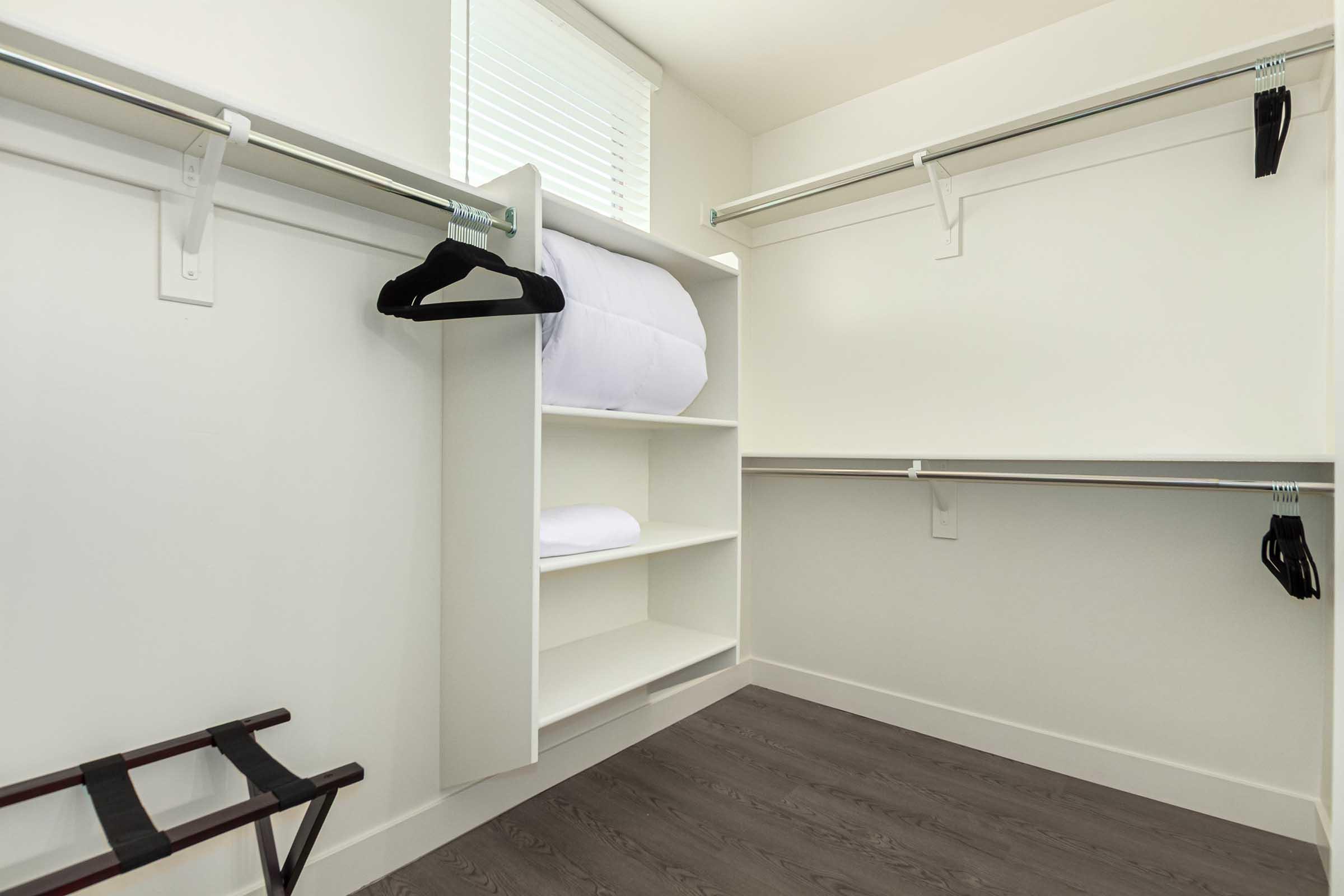 Empty closet interior featuring white walls, light wood flooring, and two hanging rods. One side has a shelf with folded bedding, while the other side is equipped with hangers. A small folding stool is positioned on the floor, and there is a window with blinds providing natural light.