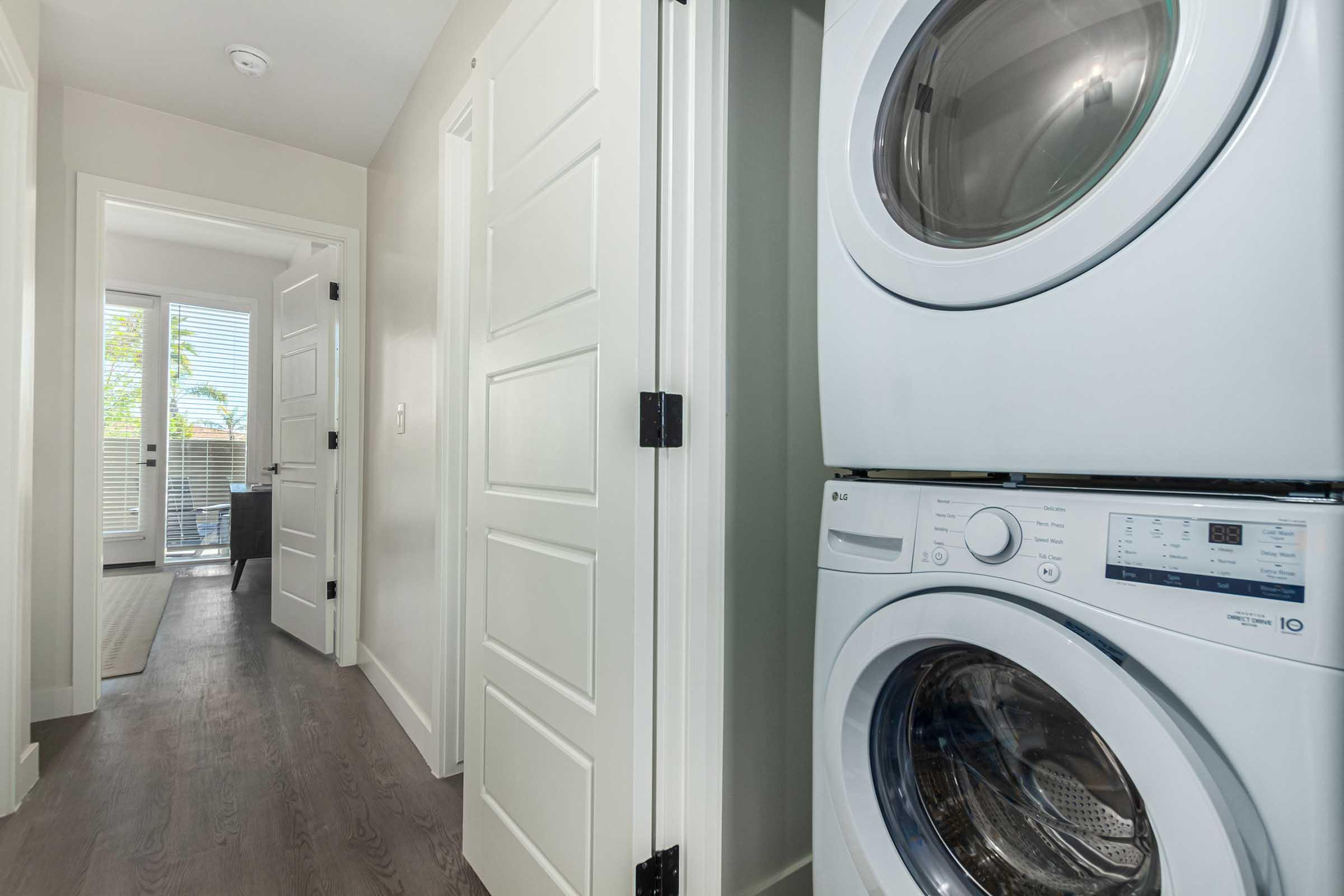 A modern laundry area featuring a stacked washer and dryer unit. The hallway is well-lit, leading to a door with a window at the end, and includes light-colored walls and wooden flooring. The overall design is clean and contemporary.