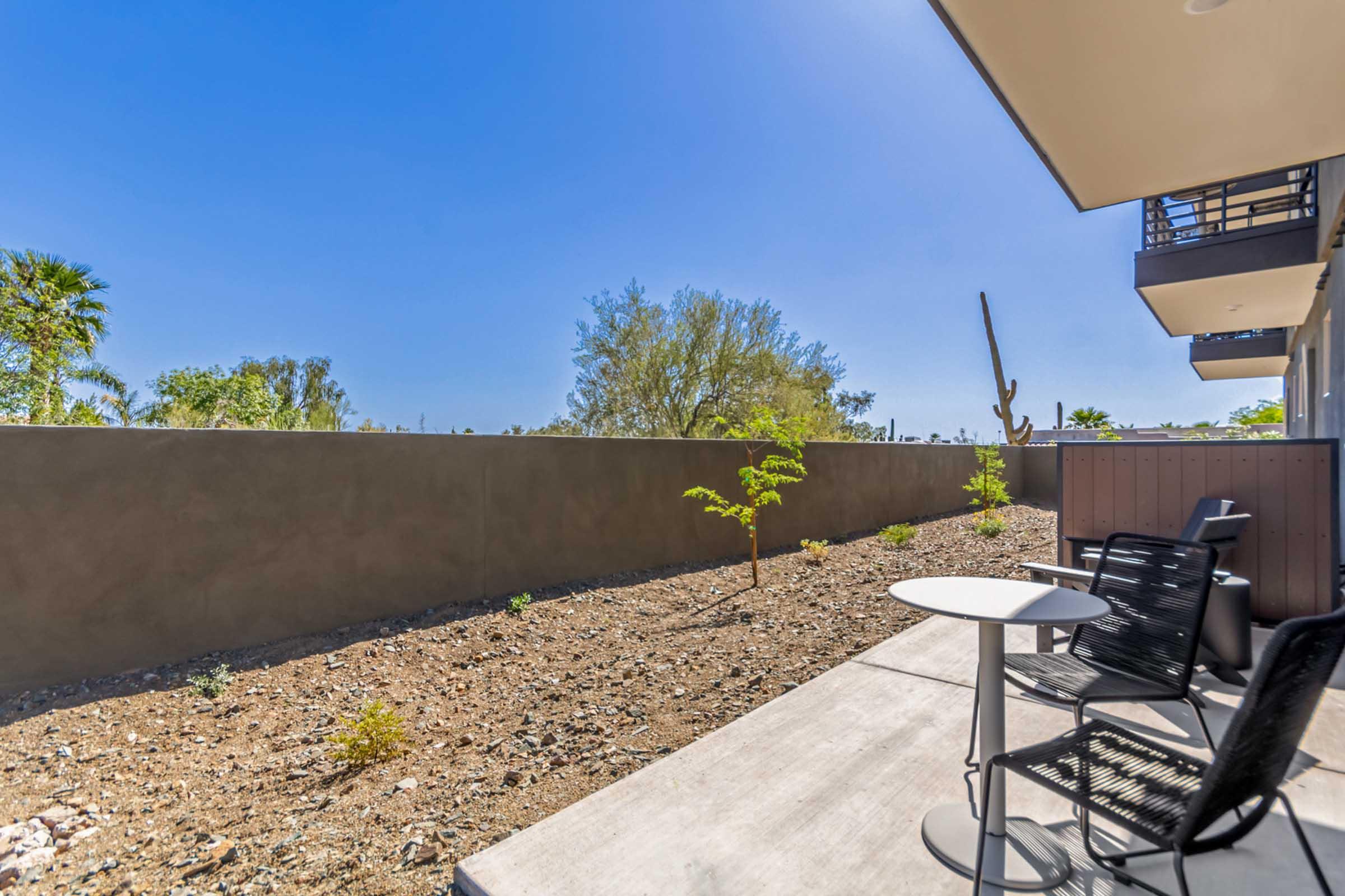 A clear blue sky above a landscaped patio area featuring two black chairs and a small round table. The ground is composed of gravel and desert plants, with a low wall in the background. Lush greenery from nearby trees adds a touch of nature to the scene.