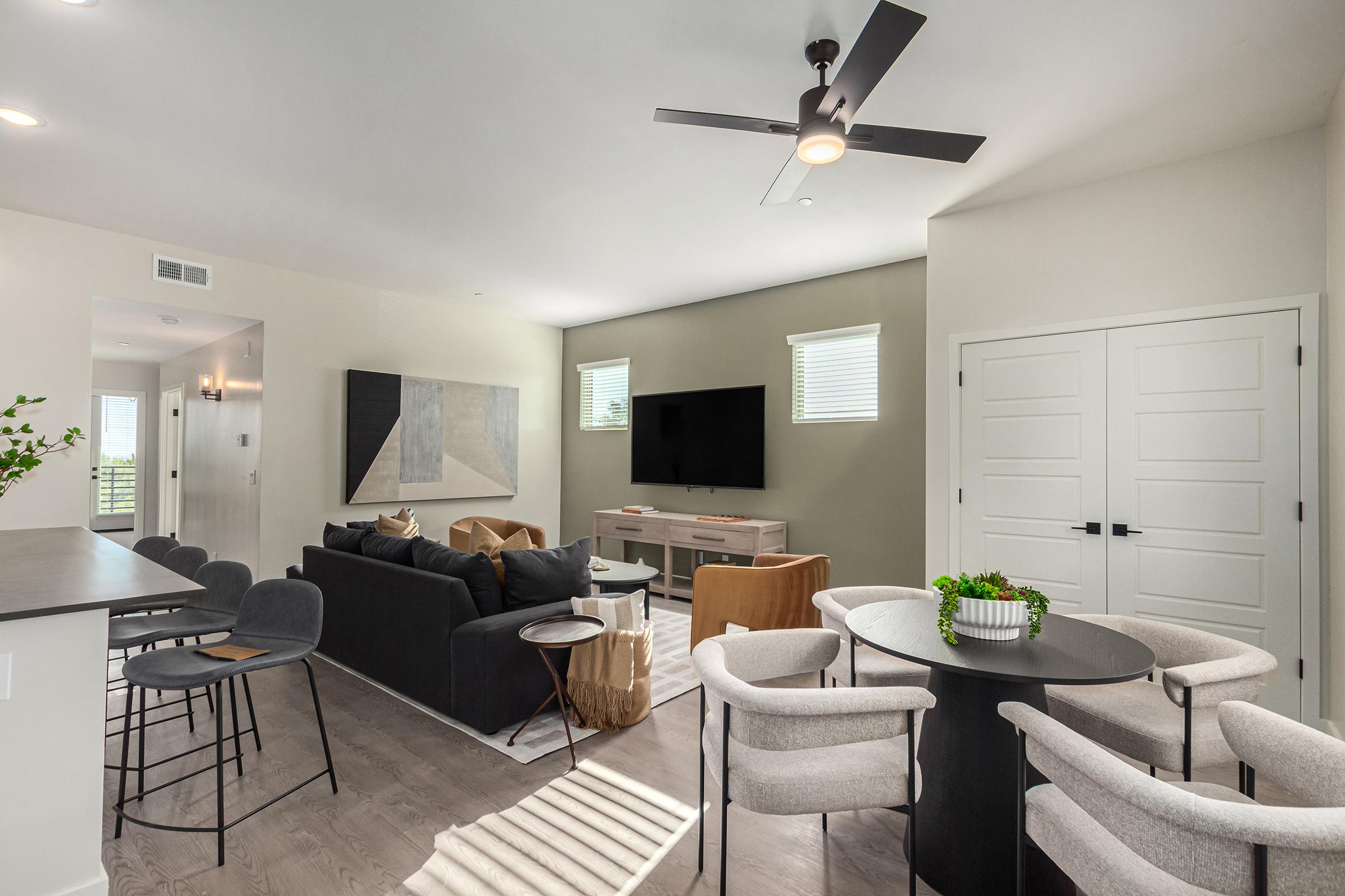 Modern living space featuring a cozy lounge area with a dark sofa, light chairs, and a coffee table. A flat-screen TV is mounted on the wall, with large windows allowing natural light. The dining area includes a round table with stylish chairs, surrounded by neutral colors and contemporary decor.
