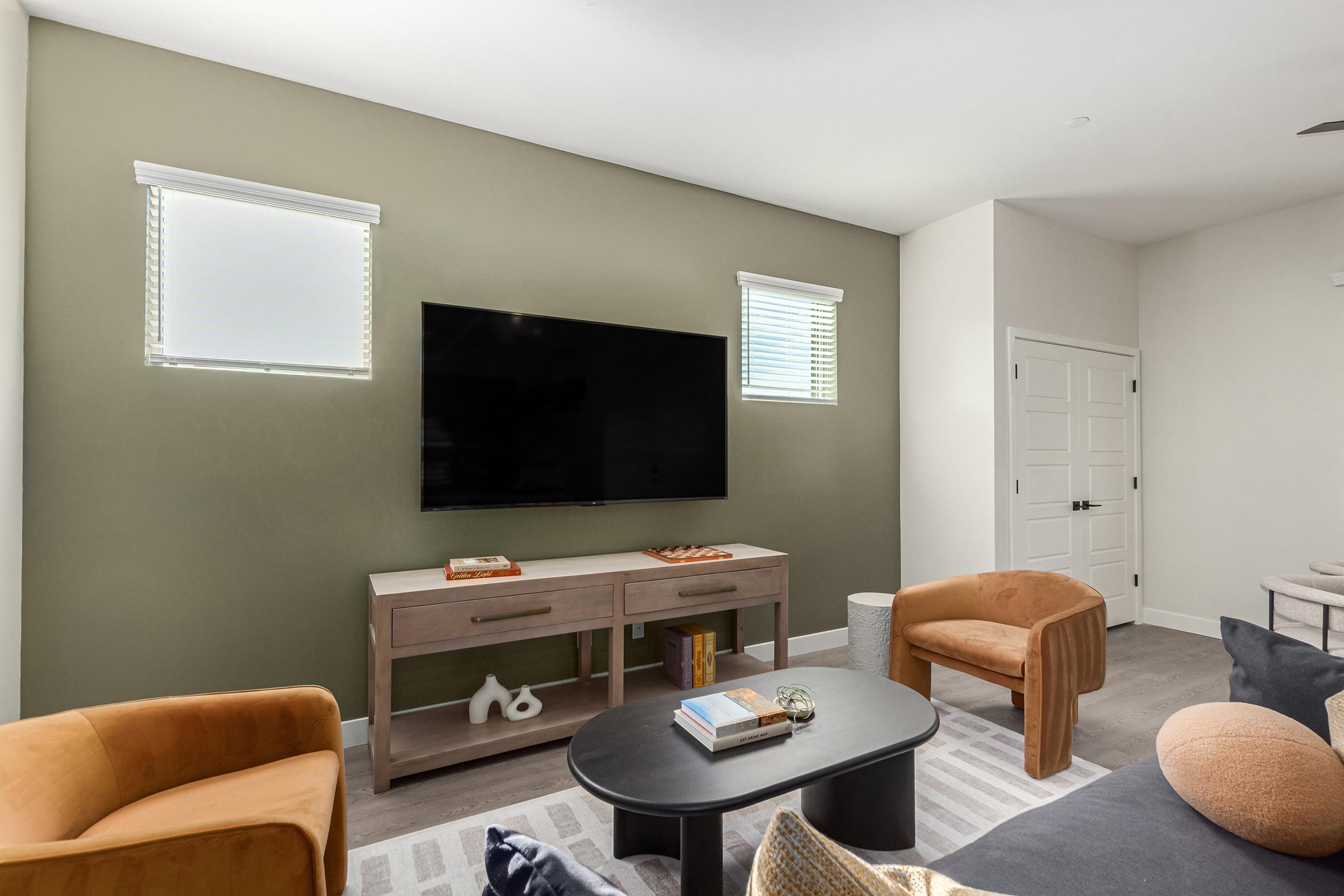 A cozy living room featuring a large flat-screen TV mounted on a green accent wall. There are two orange chairs and a round coffee table in the foreground, with a console table beneath the TV displaying decor items. Natural light enters through two windows, adding warmth to the space.