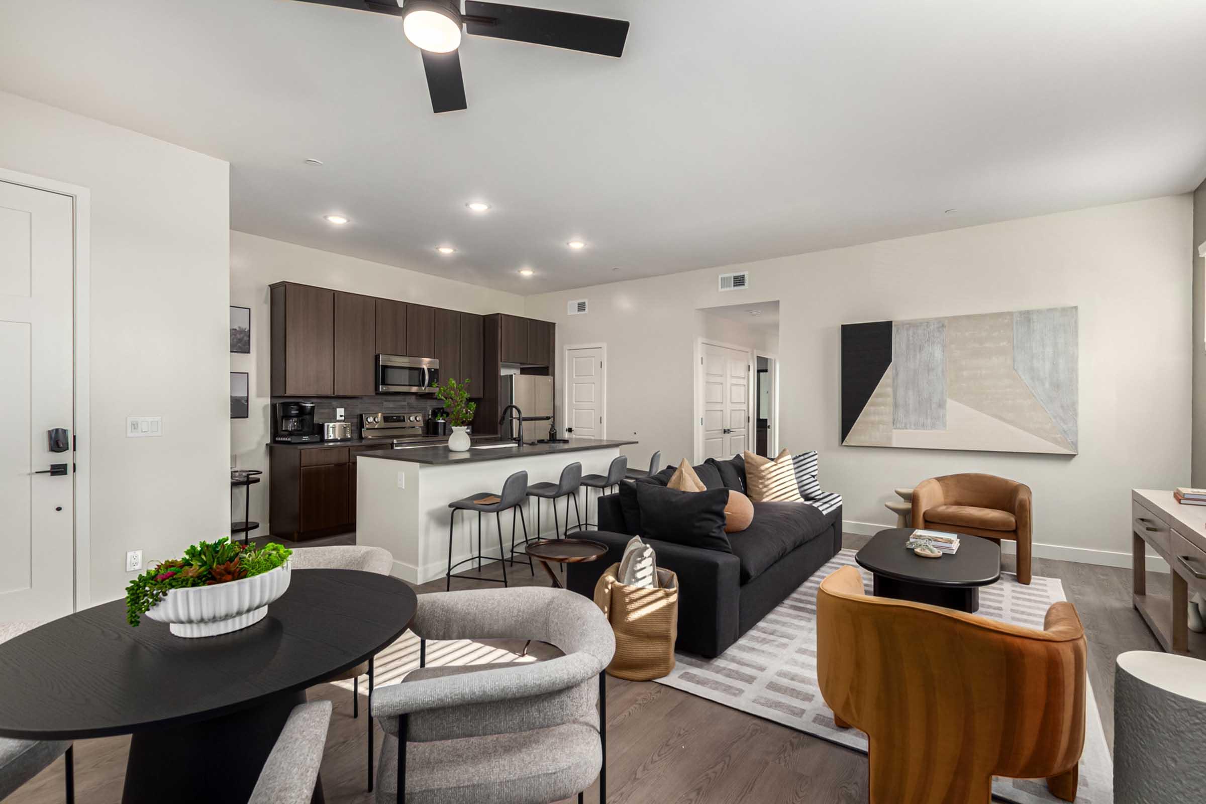 Modern living room and kitchen area with an open layout. Features a stylish dark sofa with decorative pillows, a circular wooden table, and contemporary chairs. The kitchen includes modern appliances and bar seating. Light-colored walls and wood flooring create a warm, inviting atmosphere.