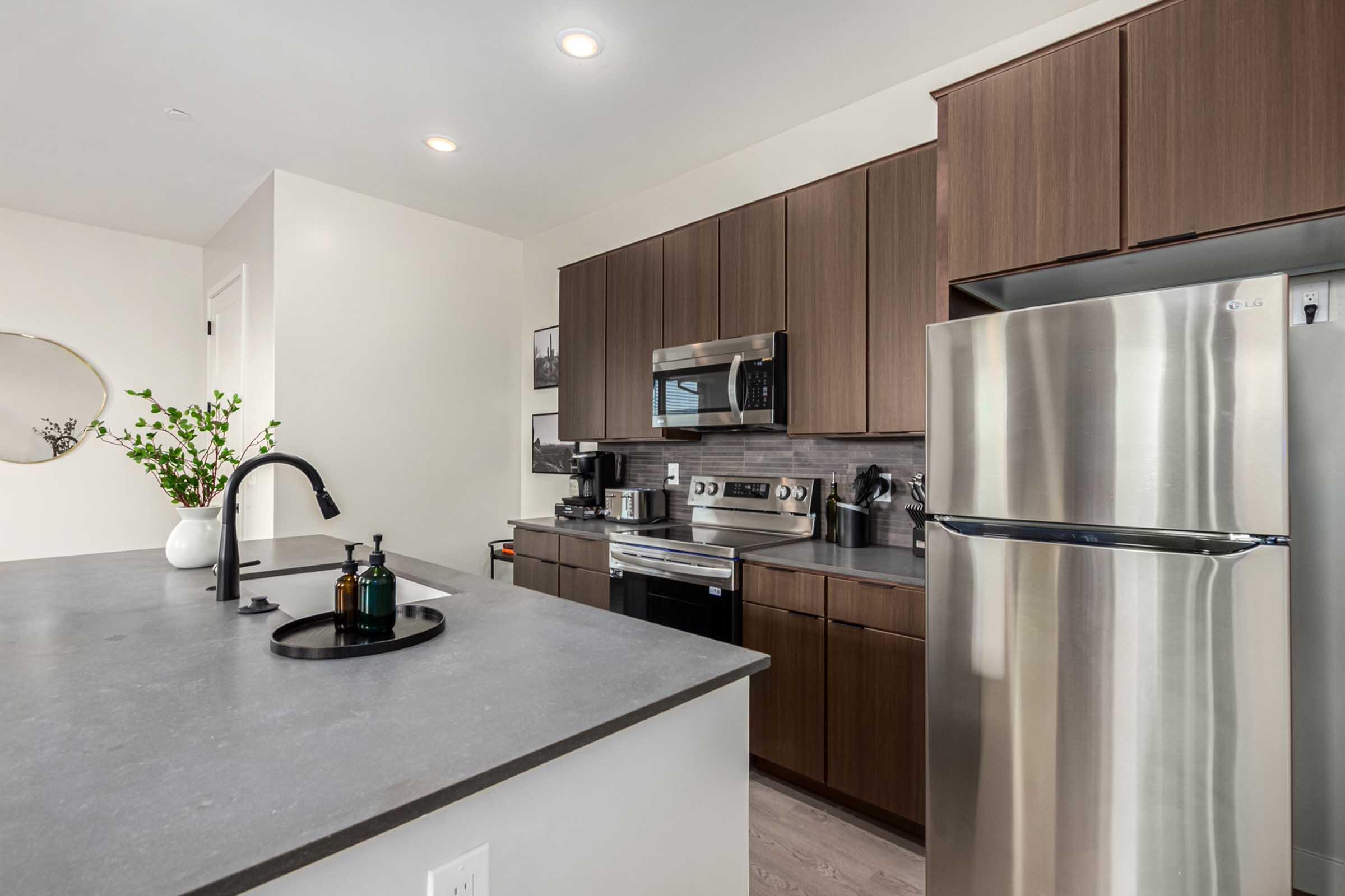 Modern kitchen featuring sleek dark wood cabinetry, stainless steel appliances including a refrigerator and stove, a spacious gray countertop with a round black tray, and a vase with greenery. Soft lighting enhances the contemporary design.