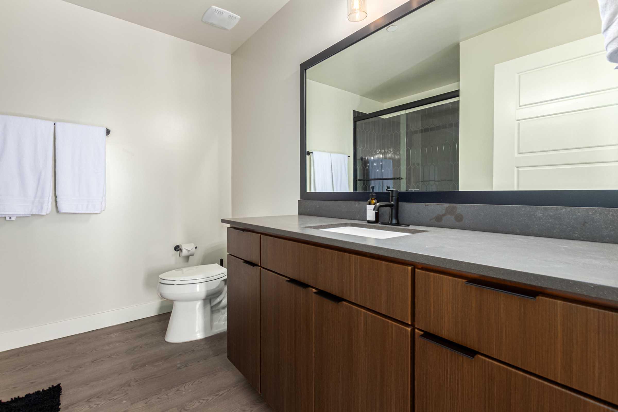 Modern bathroom featuring a sleek countertop with a sink, a large rectangular mirror, and wooden cabinetry. White towels hang on the wall, and there is a toilet beside the vanity. The flooring is a light wood tone, creating a clean and contemporary aesthetic.