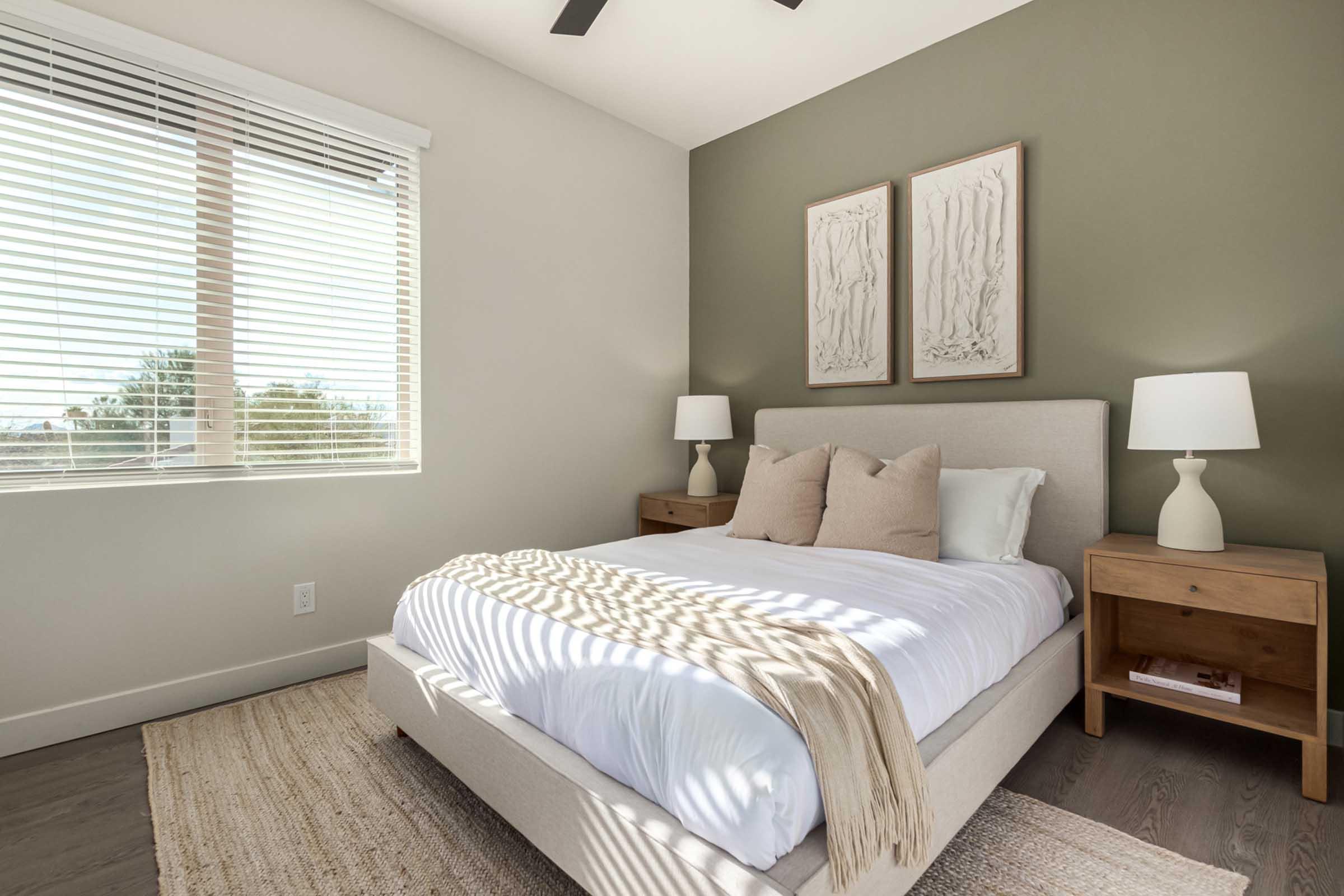 Bright and modern bedroom featuring a light grey bed with white linens and decorative pillows. There are two wooden nightstands with white lamps on either side of the bed. A window with white blinds allows natural light to fill the room, and there are botanical prints hanging on the green accent wall. A textured rug completes the cozy atmosphere.