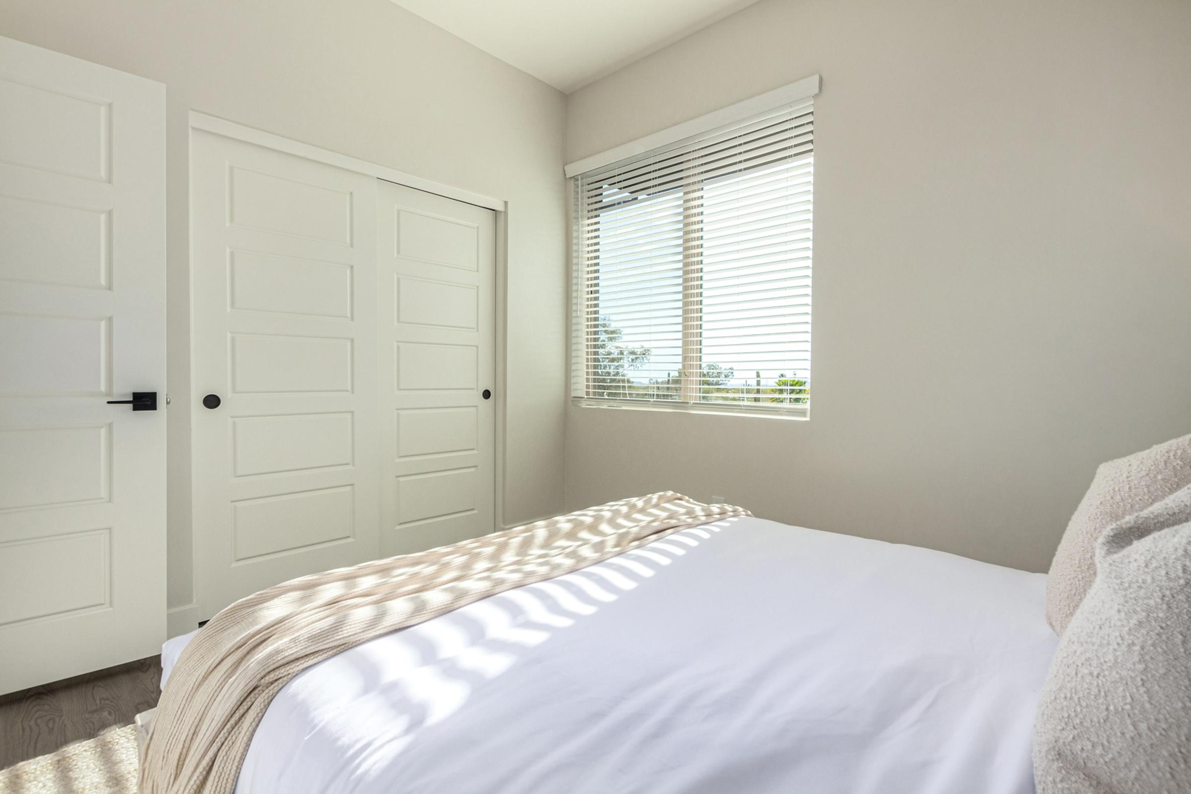 A bright, minimalistic bedroom featuring a bed with white linens, a light beige throw blanket, and sunlight casting shadows on the floor. There are white walls, a large window with blinds offering a view, and a two-door closet to the side. The overall atmosphere is clean and serene.