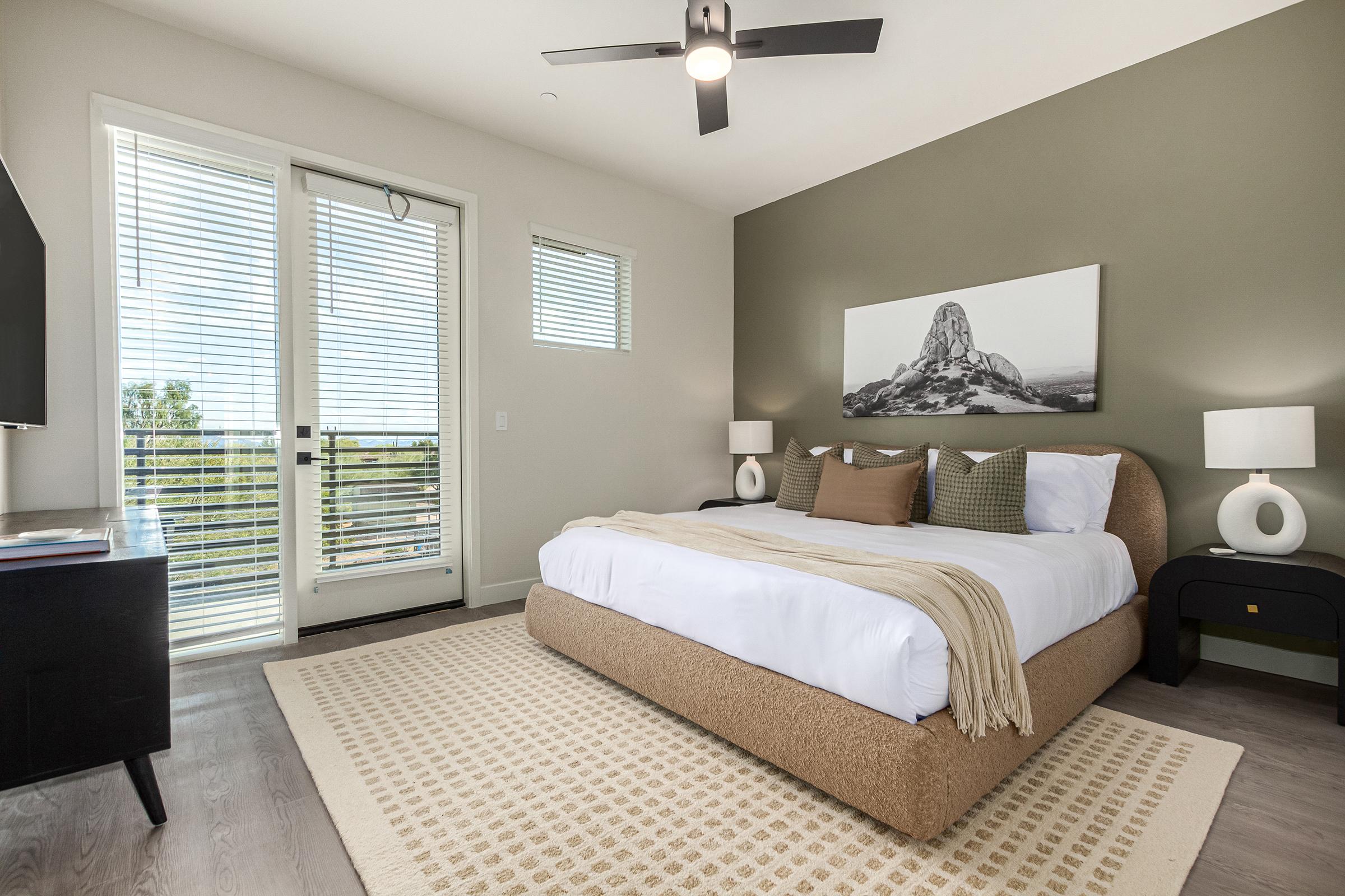 A modern bedroom featuring a large bed with a light-colored blanket, two pillows, and bedside lamps. A photo of a rock formation hangs on the wall above the bed. There are large windows with blinds allowing natural light, and a black dresser is positioned next to the bed. The room has neutral-colored walls and a light rug.