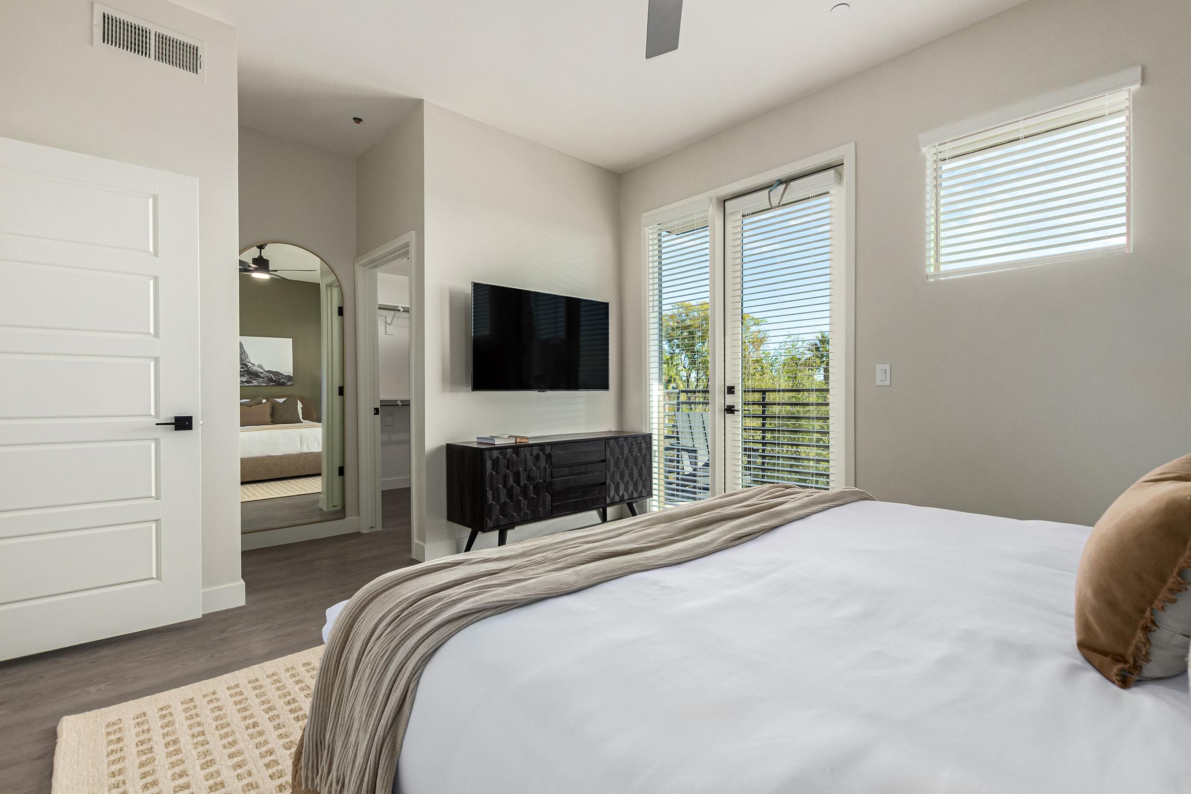 A modern bedroom featuring a neatly made bed with white linens and a decorative throw. A wall-mounted television is positioned across from the bed, and large windows with blinds allow natural light. In the background, a mirror reflects a doorway leading to another room, adding depth to the space.