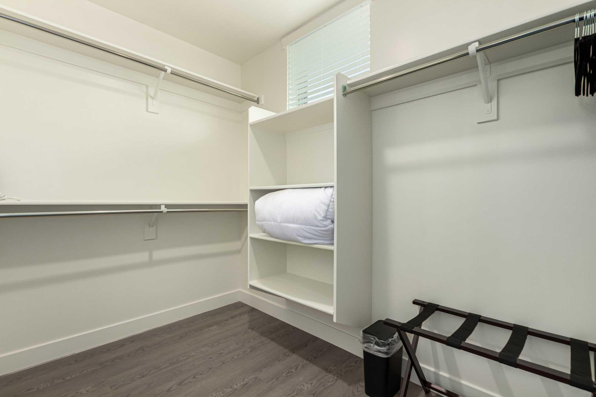 A spacious, empty closet featuring two hanging racks, a central shelving unit with a neatly folded white comforter, and a small wastebasket. The floor is a light wood finish, and natural light filters in from a window. The overall design is modern and minimalist.