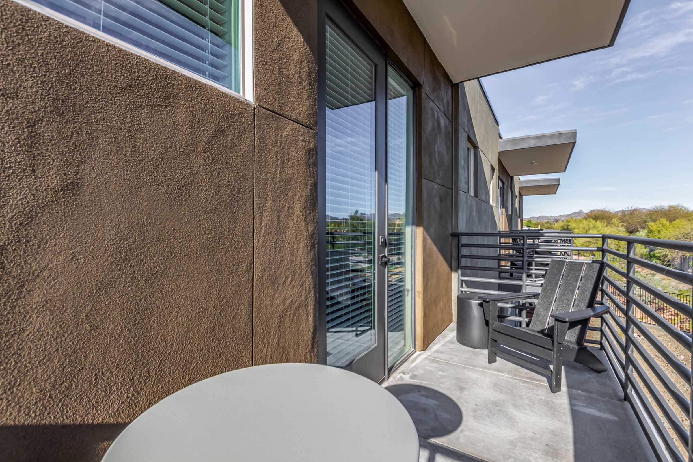 A modern balcony with a smooth gray table and a black Adirondack chair facing a railing. The glass sliding door is partially visible, flanked by large windows. Surrounding greenery and blue sky can be seen in the background, suggesting a tranquil outdoor space for relaxation.