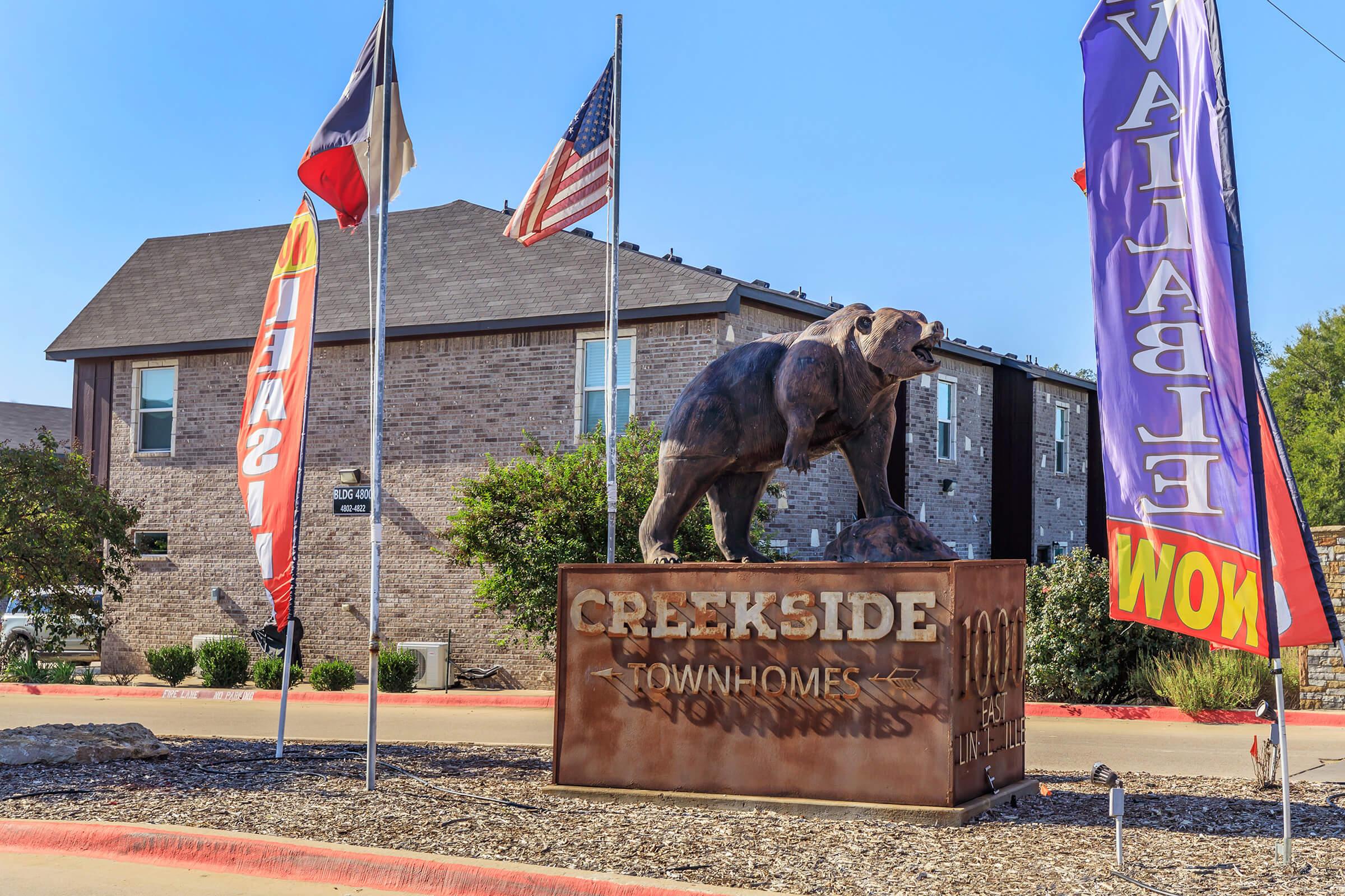 Signage for Creekside Townhomes featuring a sculpture of a bear. The area has colorful flags and an American flag displayed. The building in the background is modern in style, constructed from brick. The sign includes the text "Creekside Townhomes" and the address.