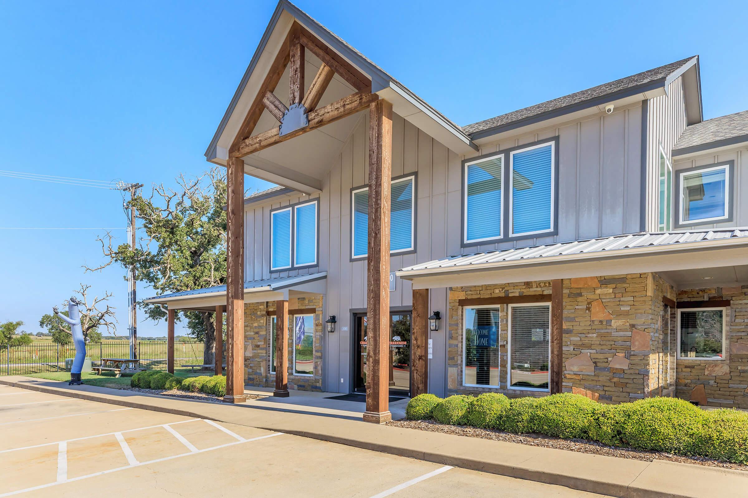 A modern building with a combination of stone and wood accents, featuring large windows and a welcoming entrance. The front area is landscaped with green bushes, and there is a parking lot visible. A tree and a statue are positioned nearby, set against a clear blue sky.