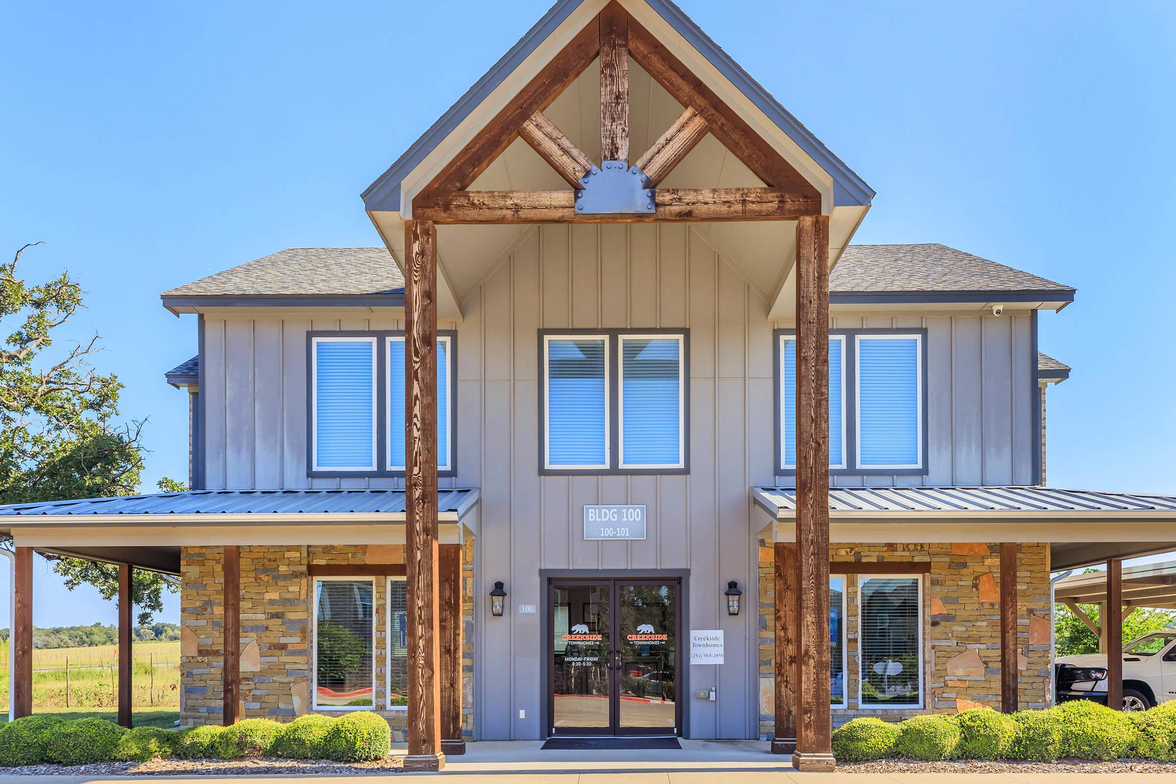 A modern building with a peaked roof and wooden accents, featuring large windows and a stone facade. The entrance is framed by two wooden columns and has a clear glass door. Lush greenery surrounds the pathway, and the background includes a clear blue sky and open fields.