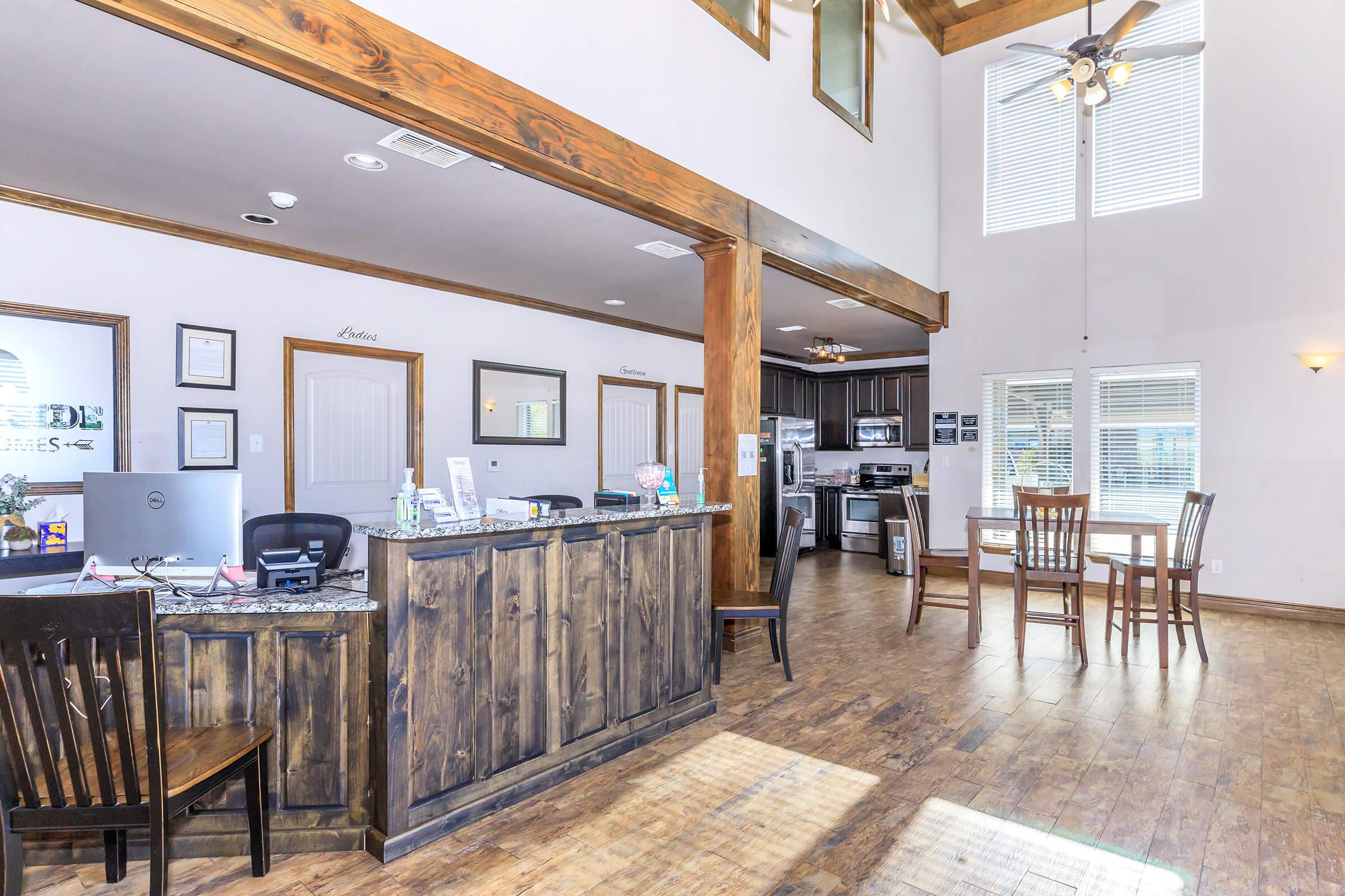 Interior of a spacious reception area featuring a wooden front desk, several chairs, and a dining table. Large windows allow natural light, highlighting the wooden beams and flooring. A kitchen area is visible in the background, with equipment and cabinets. The space is well-organized and inviting.
